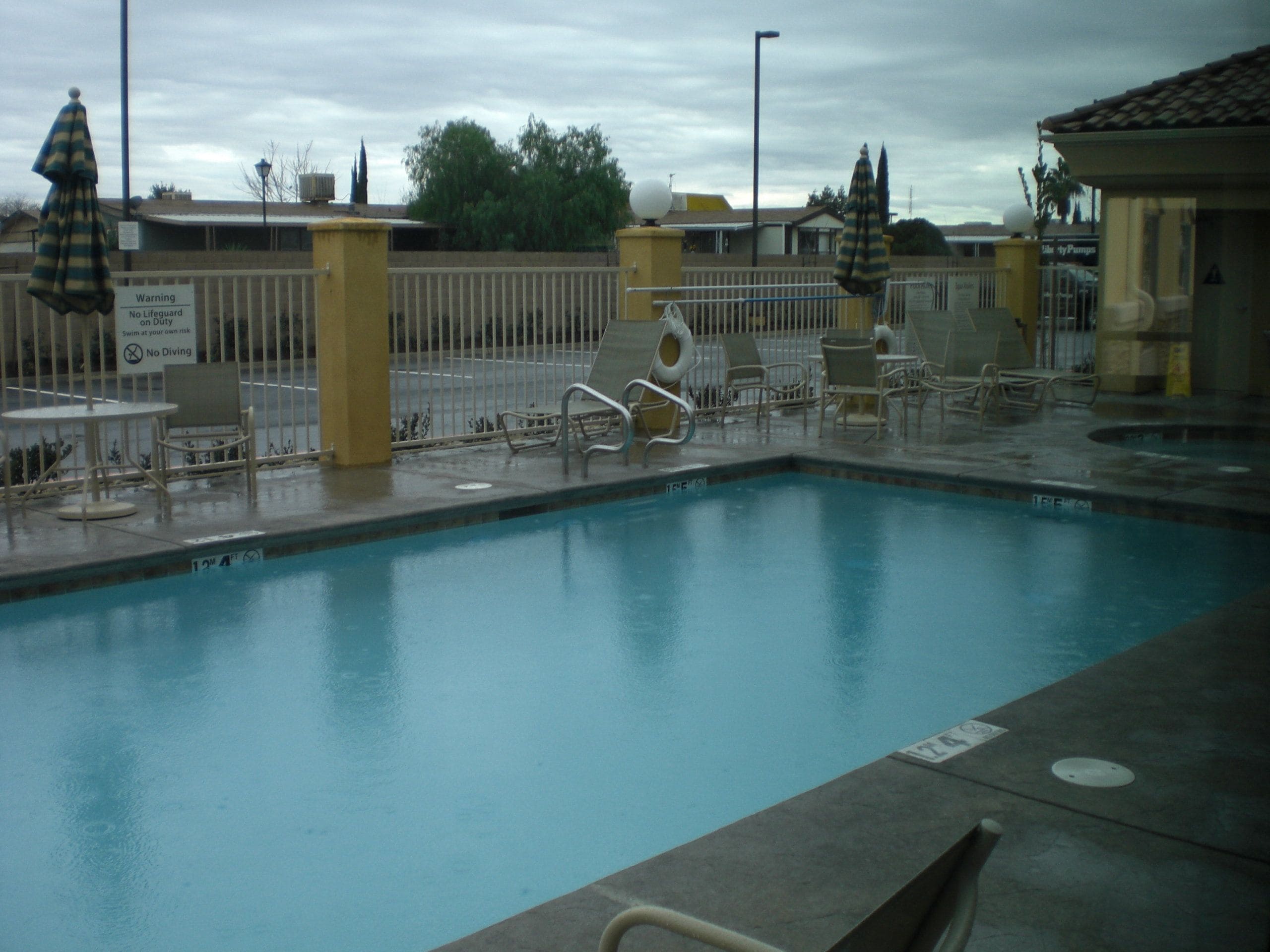 A swimming pool with lounge chairs and umbrellas, surrounded by a fence and a lifeguard warning sign.