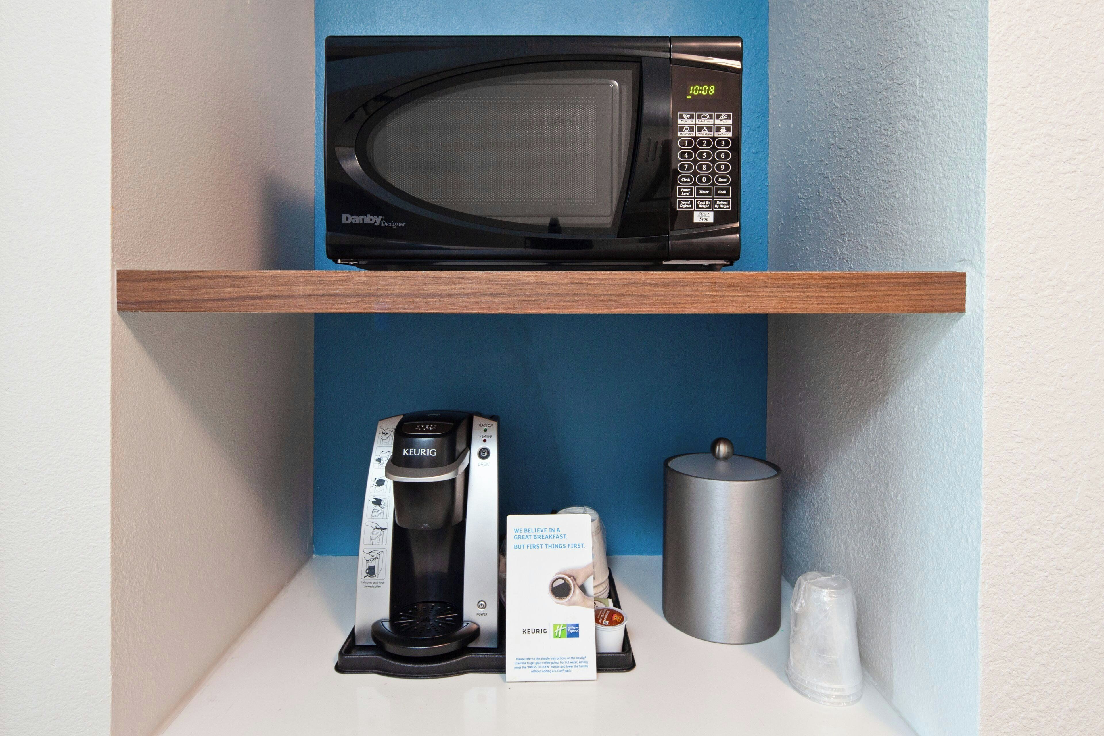 A microwave on a shelf, a Keurig coffee maker, a glass, and a stainless steel container.