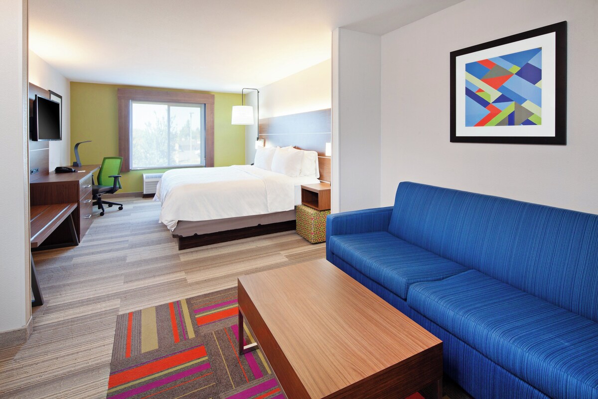 A hotel room with a bed, a blue sofa, a desk with a chair, a television, and a framed picture on the wall.