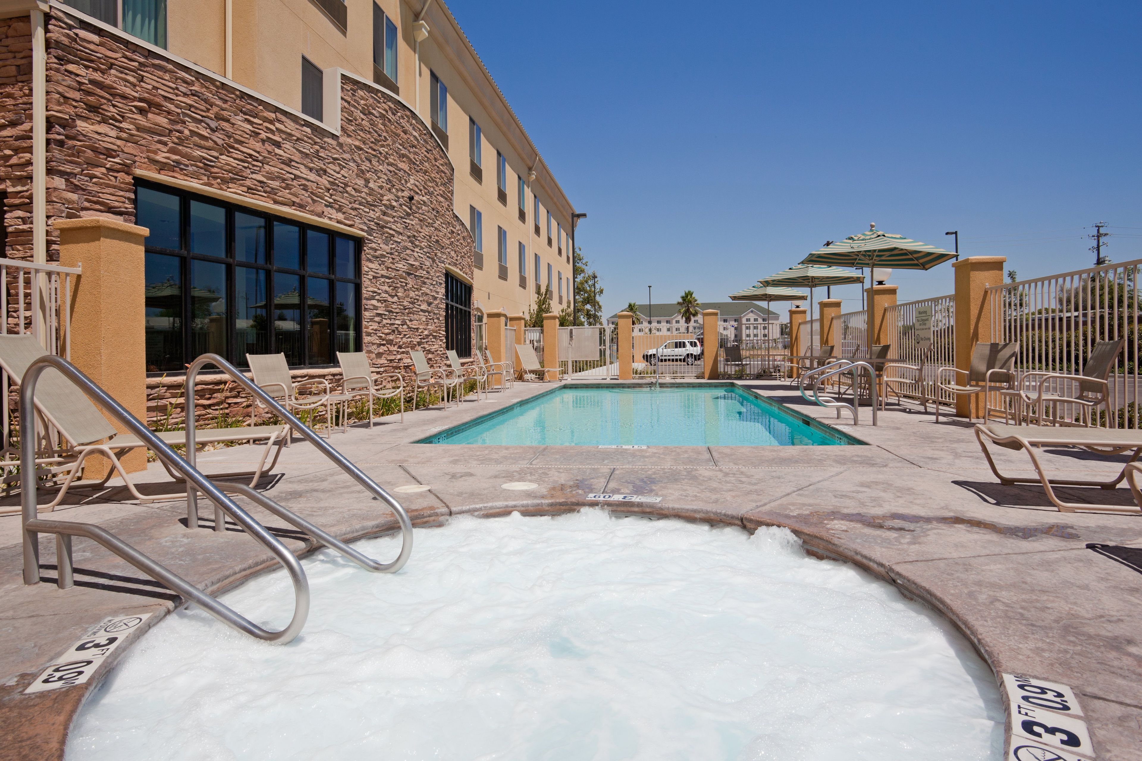 A pool area with lounge chairs and a hot tub, surrounded by a stone building with large windows.