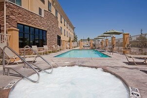 Zona de piscina con sillas de descanso y un jacuzzi, rodeada por un edificio de piedra con amplias ventanas.