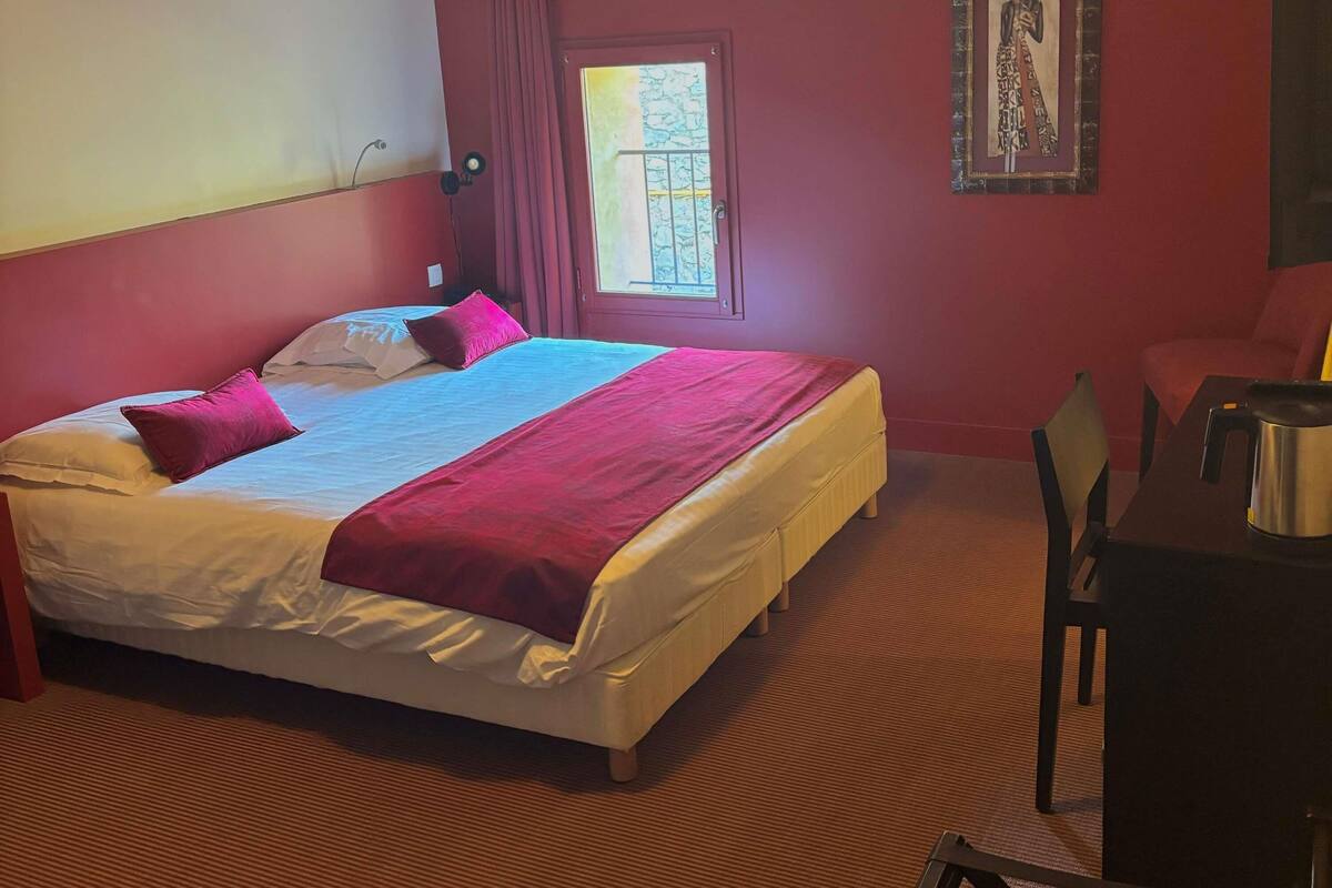 Une chambre d’hôtel avec un lit, un bureau, une chaise, une fenêtre et un tableau encadré au mur.