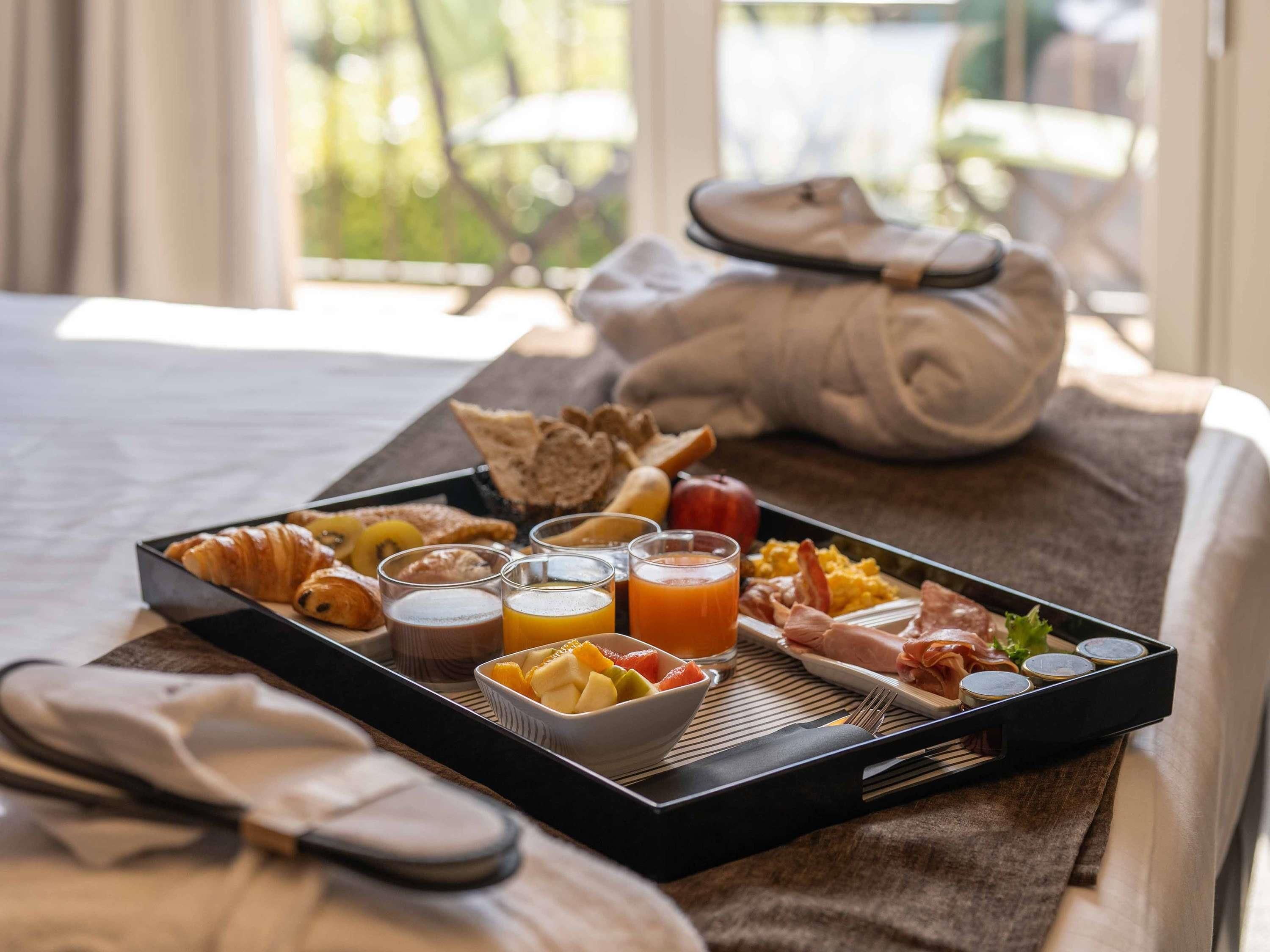 Un plateau de petit-déjeuner avec des croissants, des fruits, des œufs brouillés, du bacon et du jus, posé sur un lit.