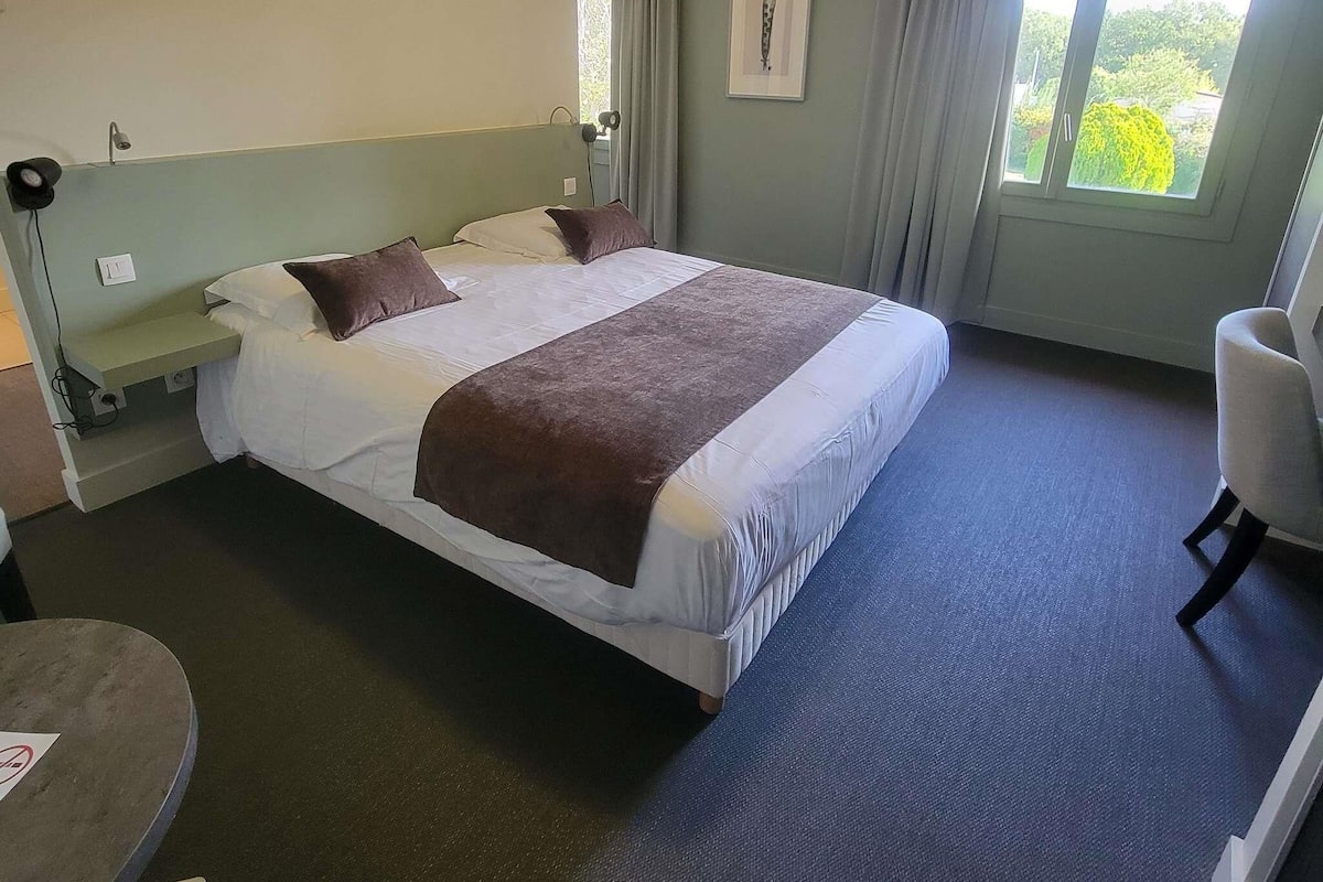 Ein Hotelzimmer mit Bett, Stuhl, Schreibtisch, einem Fenster mit Vorhängen und einem Bild an der Wand.