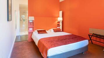 Habitación de hotel con una cama grande, un banco, zona de ducha y una lámpara de pared.