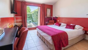 A hotel room with a bed, a desk, a chair, a window with curtains, and a view of greenery outside. - Hotel Mercure Aix en Provence Sainte Victoire (Chateauneuf-le-Rouge)