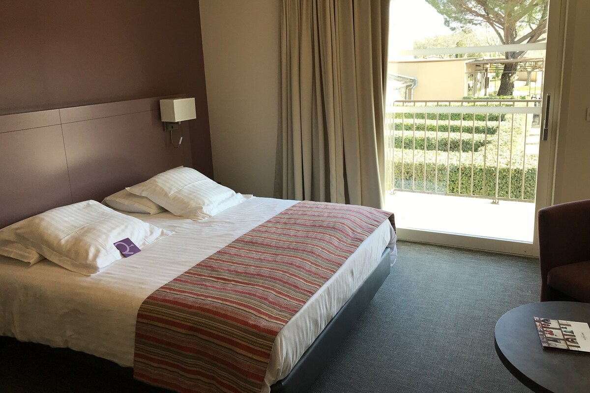 Quarto de hotel com cama, mesa de cabeceira, uma cadeira e vista para uma varanda com vegetação.