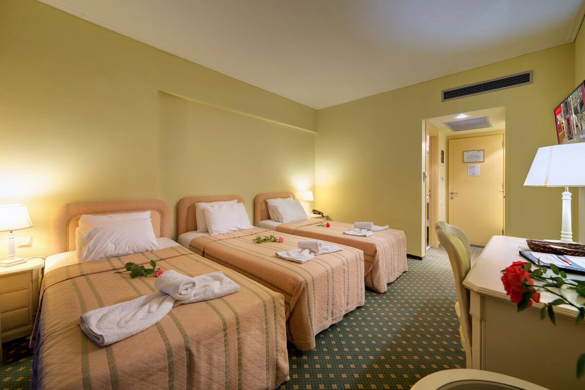 Une chambre d’hôtel avec trois lits, un bureau et une chaise. Il y a des serviettes sur les lits et une rose sur le bureau.