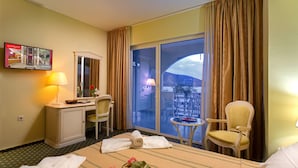 Habitación de hotel con cama, escritorio, silla, mesita, televisor y balcón con cortinas.