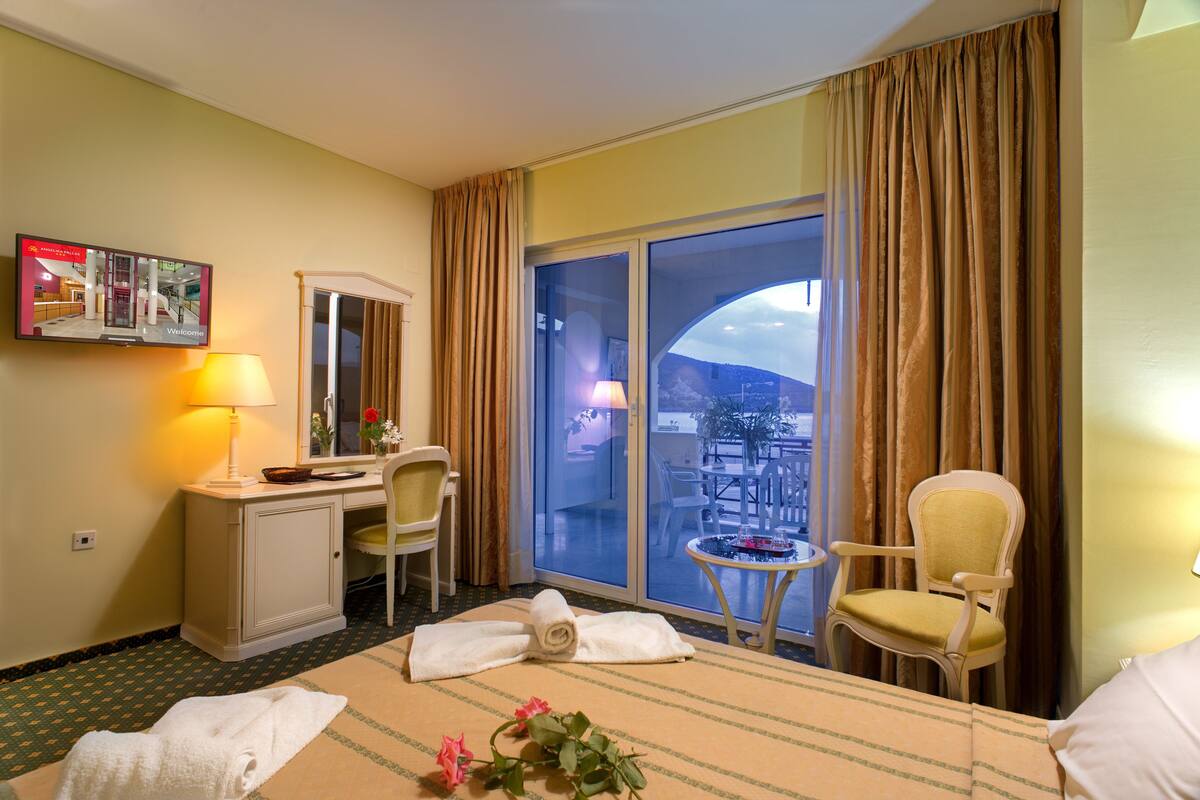 Une chambre d’hôtel comprenant un lit, un bureau, une chaise, une petite table, un téléviseur et un balcon avec des rideaux.