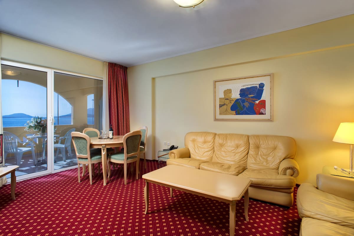Une chambre d’hôtel avec une moquette rouge à motifs, un canapé beige, une table à manger avec des chaises, une grande fenêtre donnant sur un beau paysage et une œuvre d’art encadrée accrochée au mur.