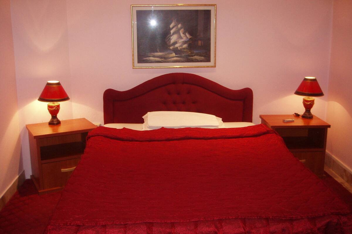 Una cama con cabecera roja, dos mesitas de noche con lámparas, un cuadro en la pared y una colcha roja.