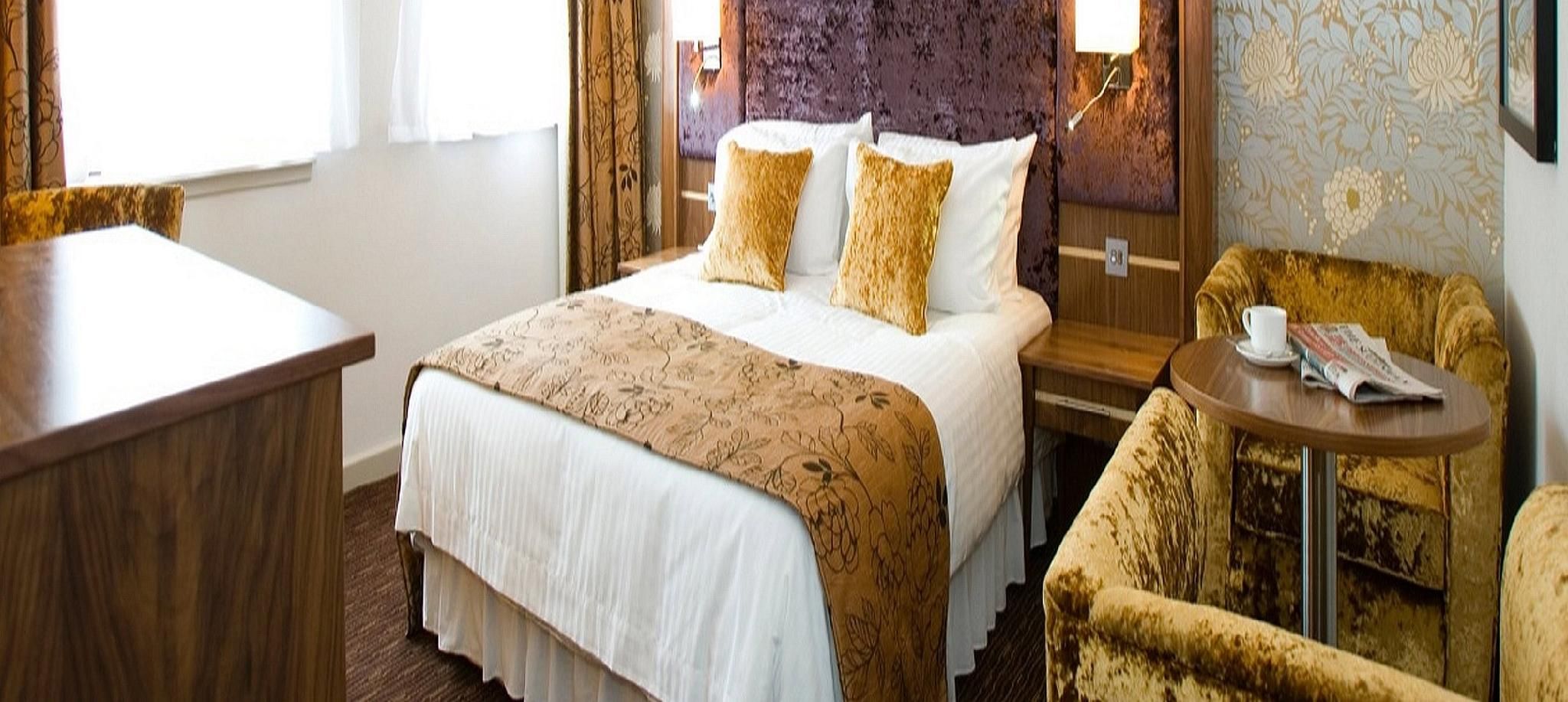 A hotel room with a bed, a desk, a chair, and a small table with a newspaper.