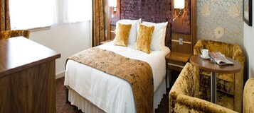 A hotel room with a bed, a desk, a chair, and a small table with a newspaper.