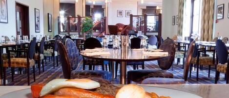 Comedor de hotel con mesas redondas, sillas y una mesa de buffet con alimentos.