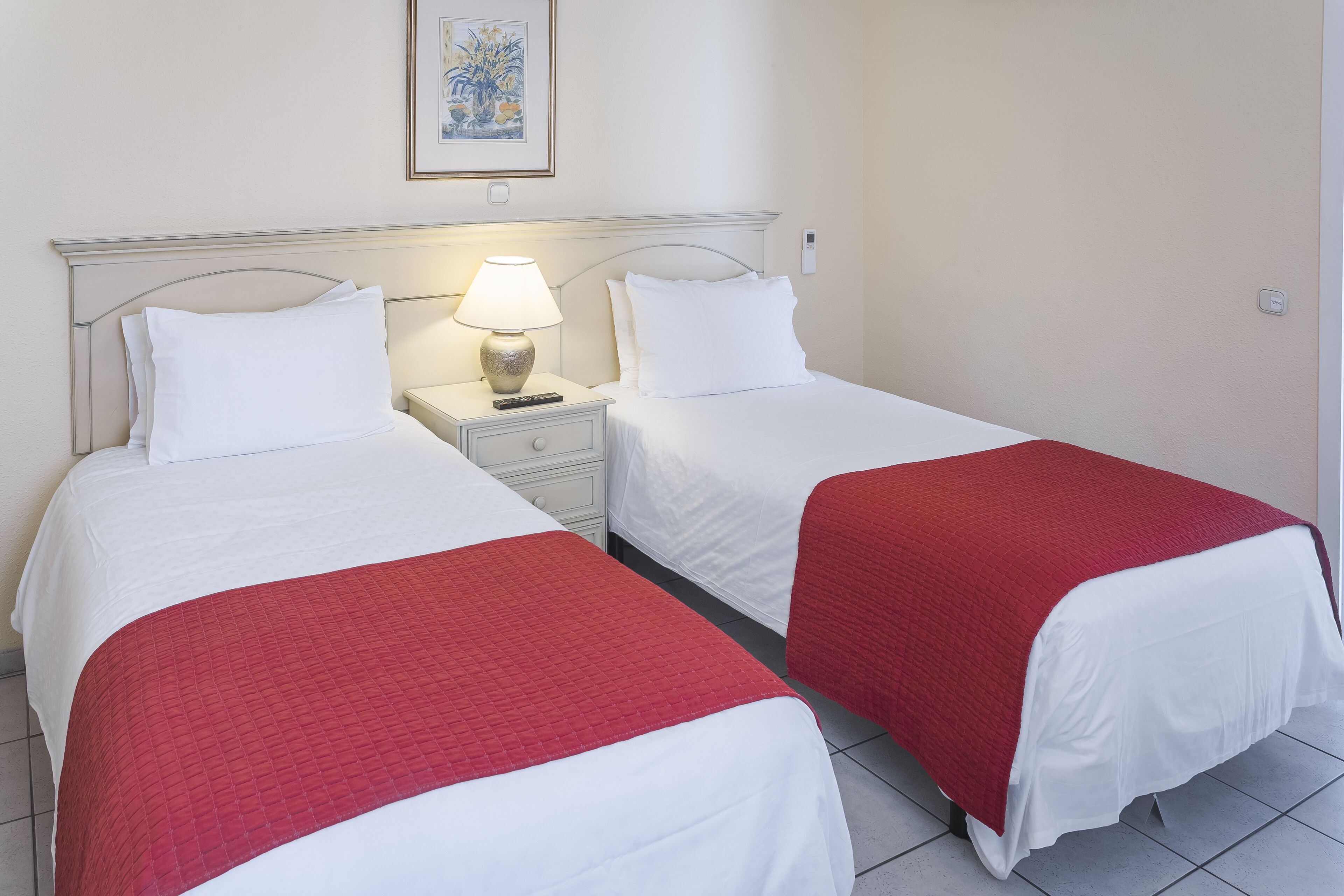 Ein Hotelzimmer mit zwei Betten, beide mit weißer Bettwäsche und roten Zudecken. Dazwischen befindet sich ein Nachttisch mit einer Lampe.