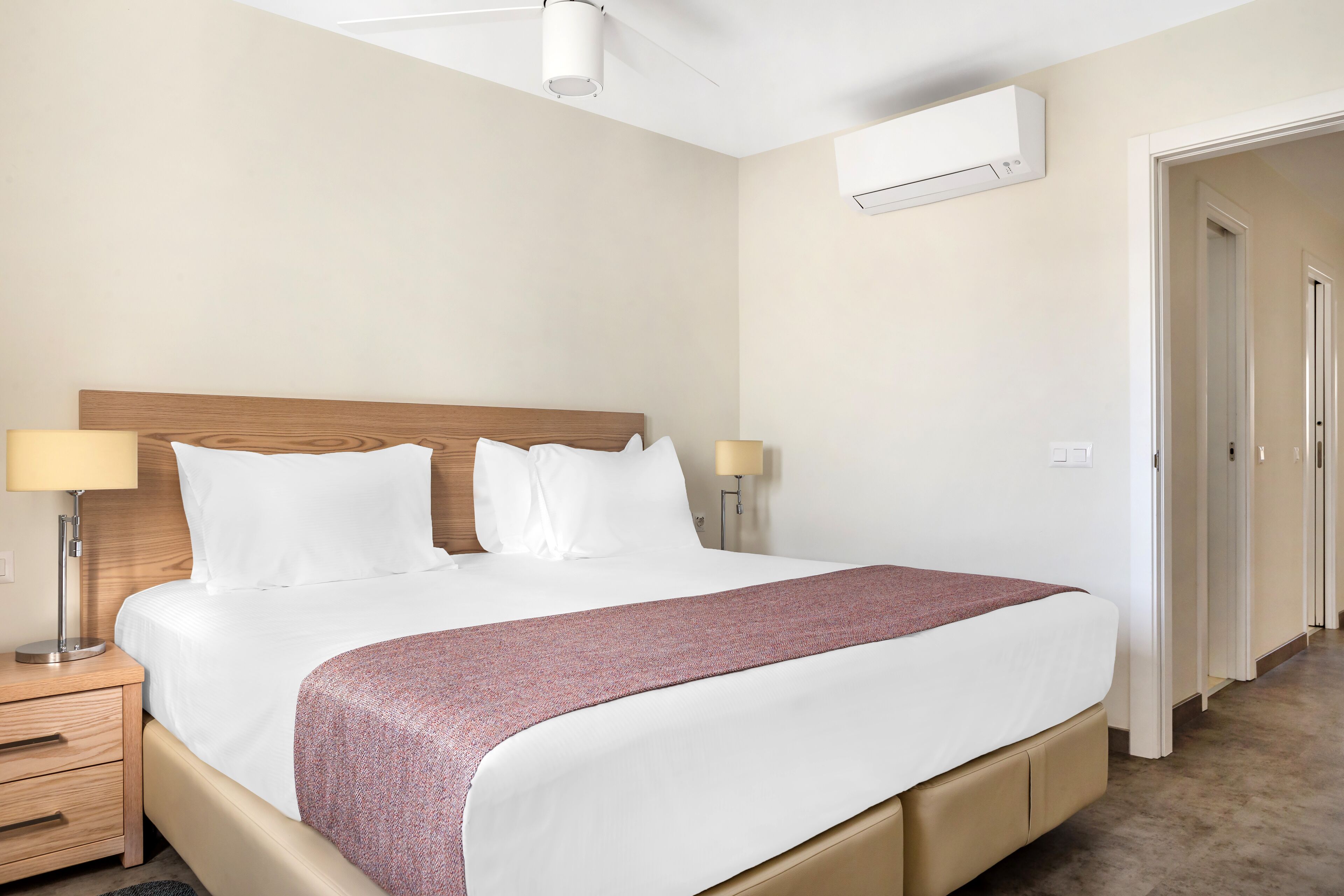 A hotel room with a bed, bedside lamps, a ceiling fan, an air conditioning unit, and a door leading to another room.