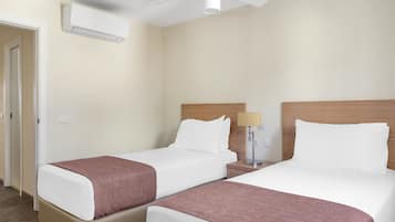 A hotel room with two beds, a ceiling fan, an air conditioning unit, and a bedside table with a lamp.