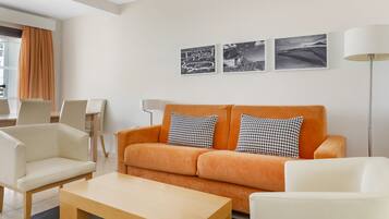Een moderne woonkamer met een bank, twee fauteuils, een salontafel en een vloerlamp. Drie zwart-witfoto's hangen aan de muur.