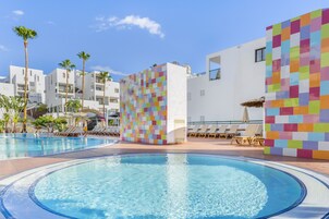 Una piscina con sillas de descanso y palmeras, rodeada de edificios blancos con paredes de mosaicos de colores.