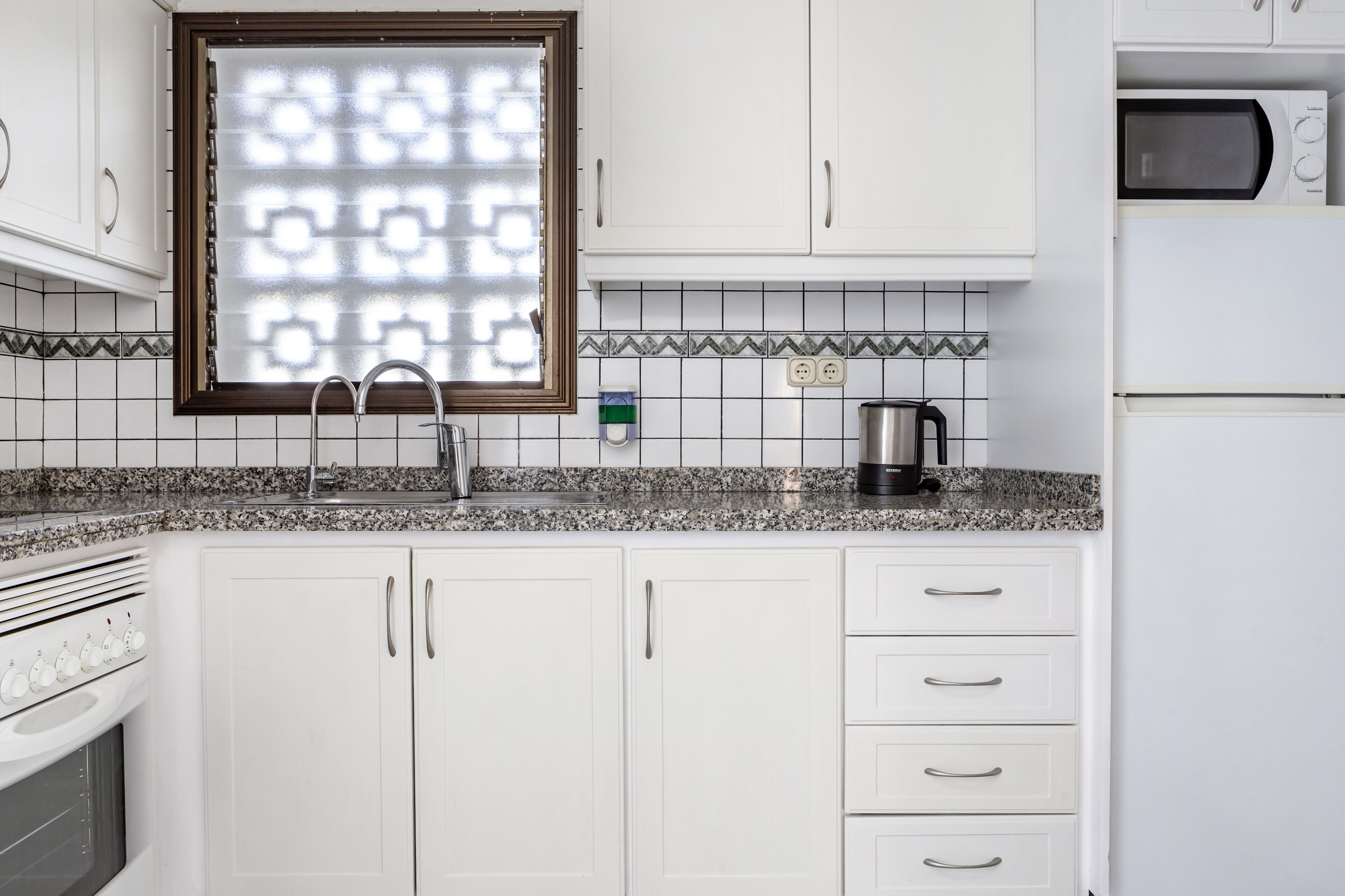Een keuken met witte kastjes, een granieten aanrecht, een roestvrijstalen spoelbak en een raam met een decoratief jaloezie, plus een magnetron.