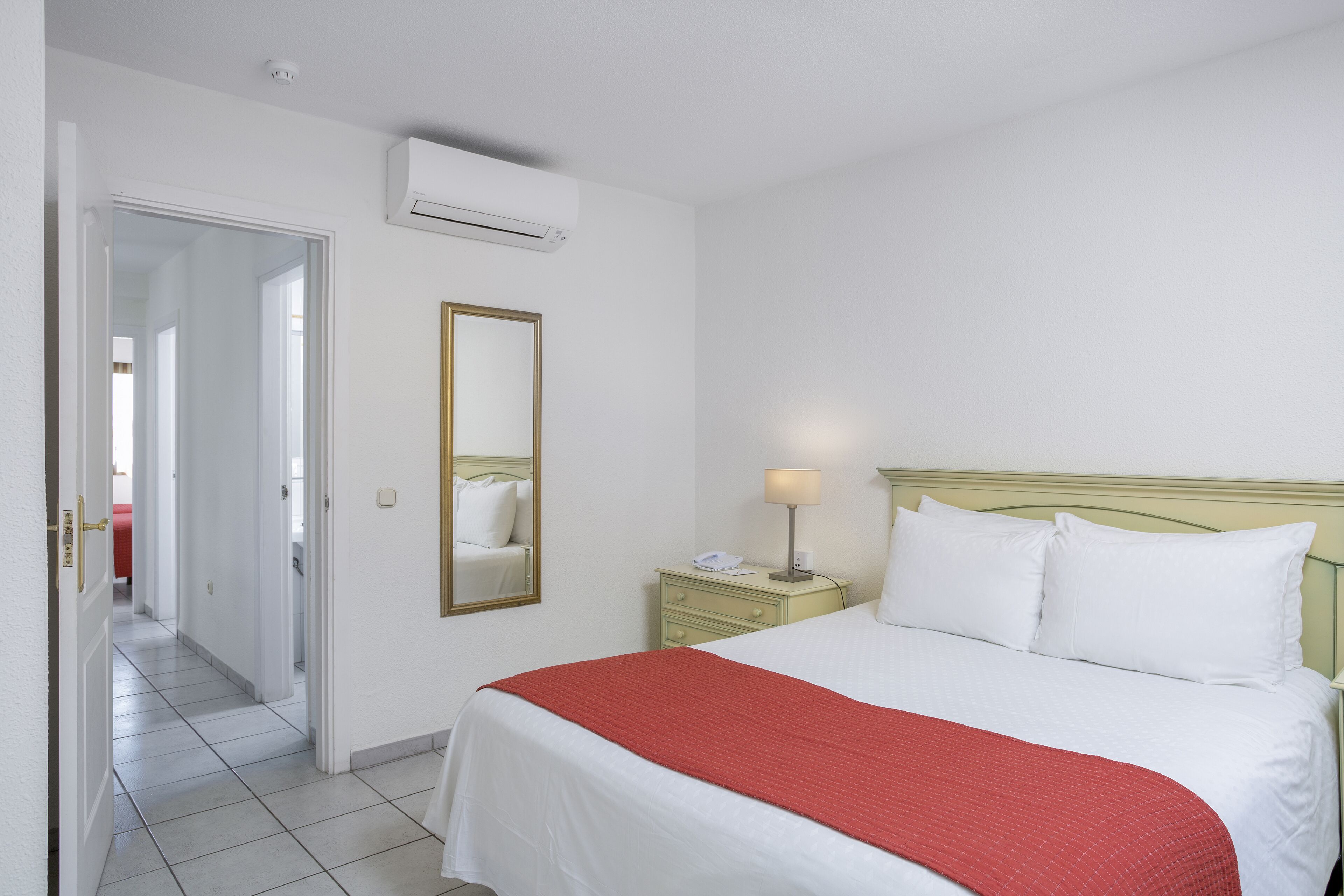 Una habitación de hotel con una cama, una mesita de noche con lámpara, un espejo y un equipo de aire acondicionado.