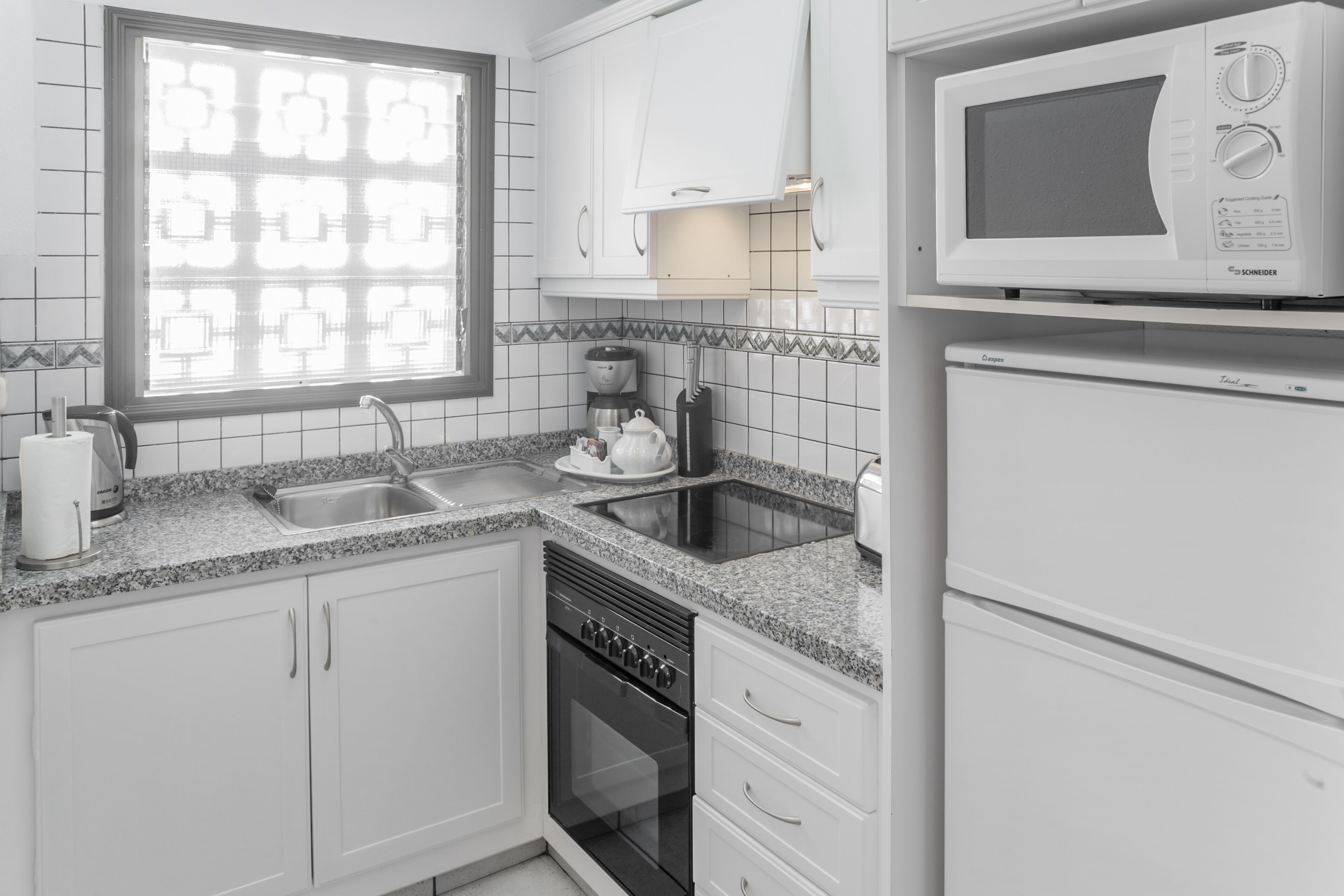 A modern kitchen with white cabinets, a granite countertop, a built-in oven, a microwave, and a window with blinds.