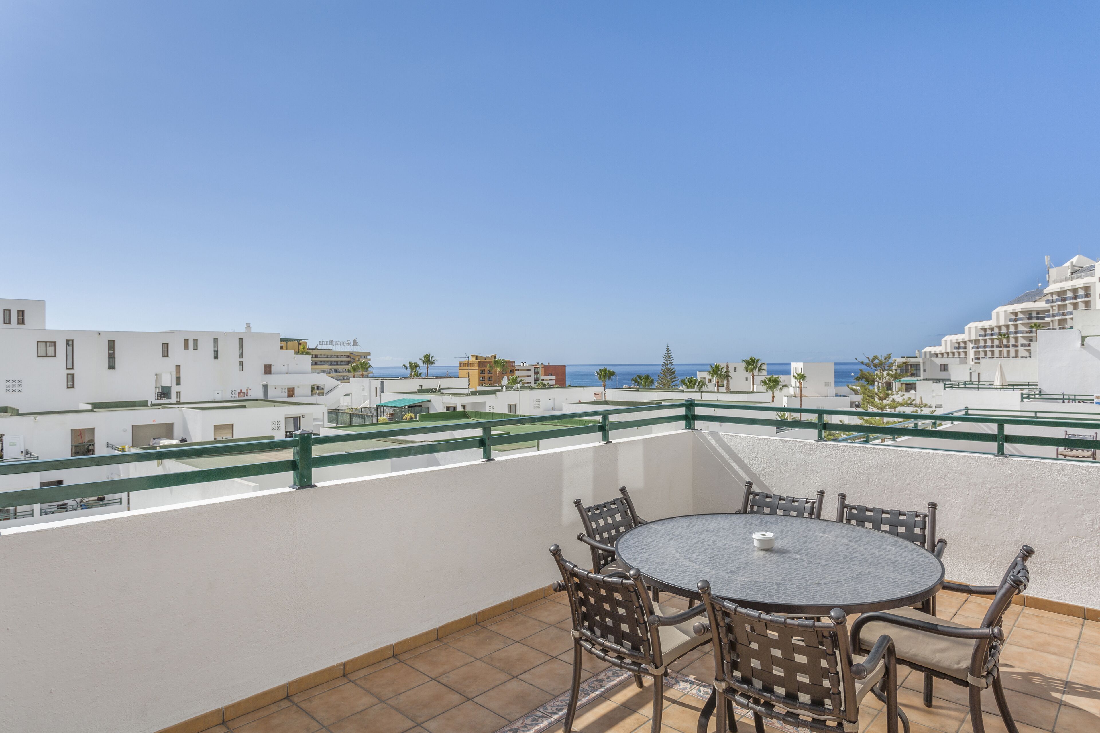 Una terraza en la azotea con una mesa redonda y sillas, que ofrece vistas al mar y edificios circundantes.