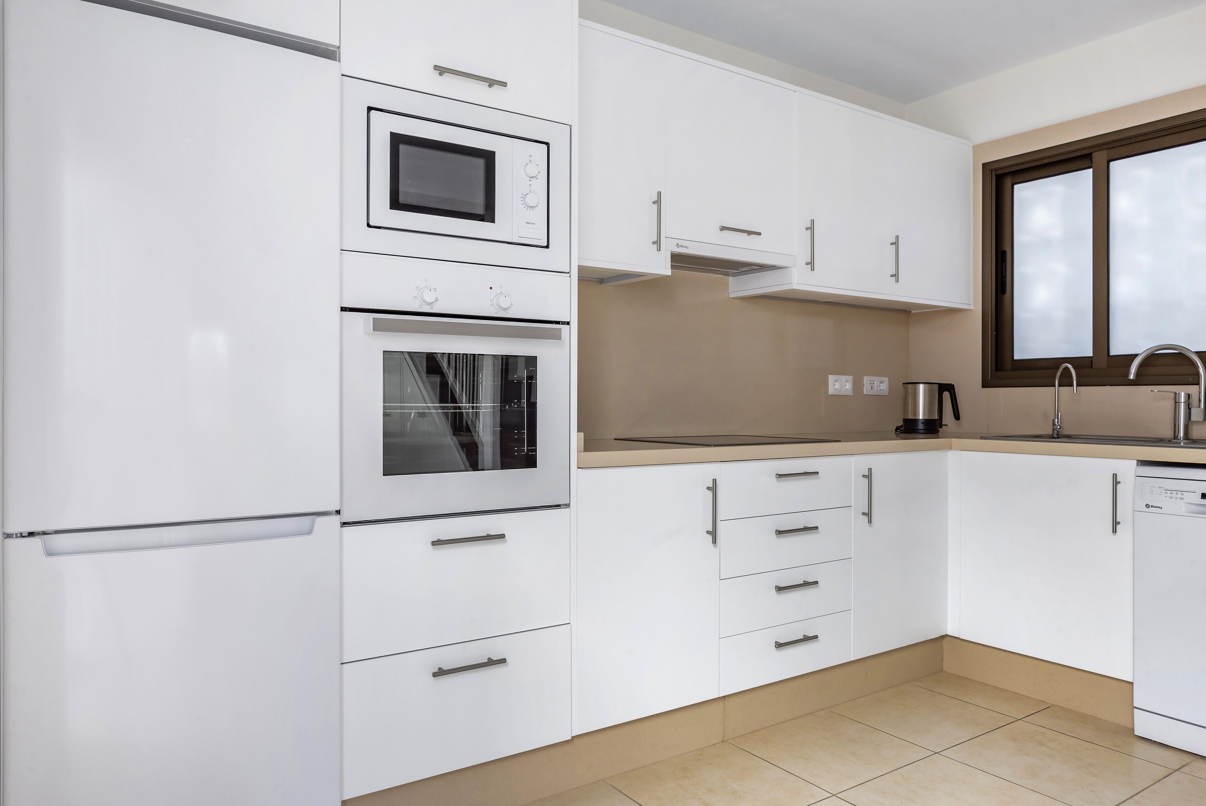 A modern kitchen with white cabinets, a built-in oven, a microwave, and a window above the sink.