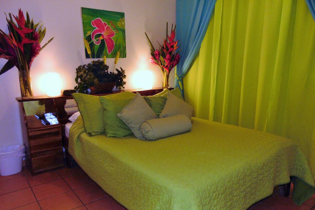 A bed with a green bedspread, a nightstand with a lamp, and a painting of a flower on the wall.