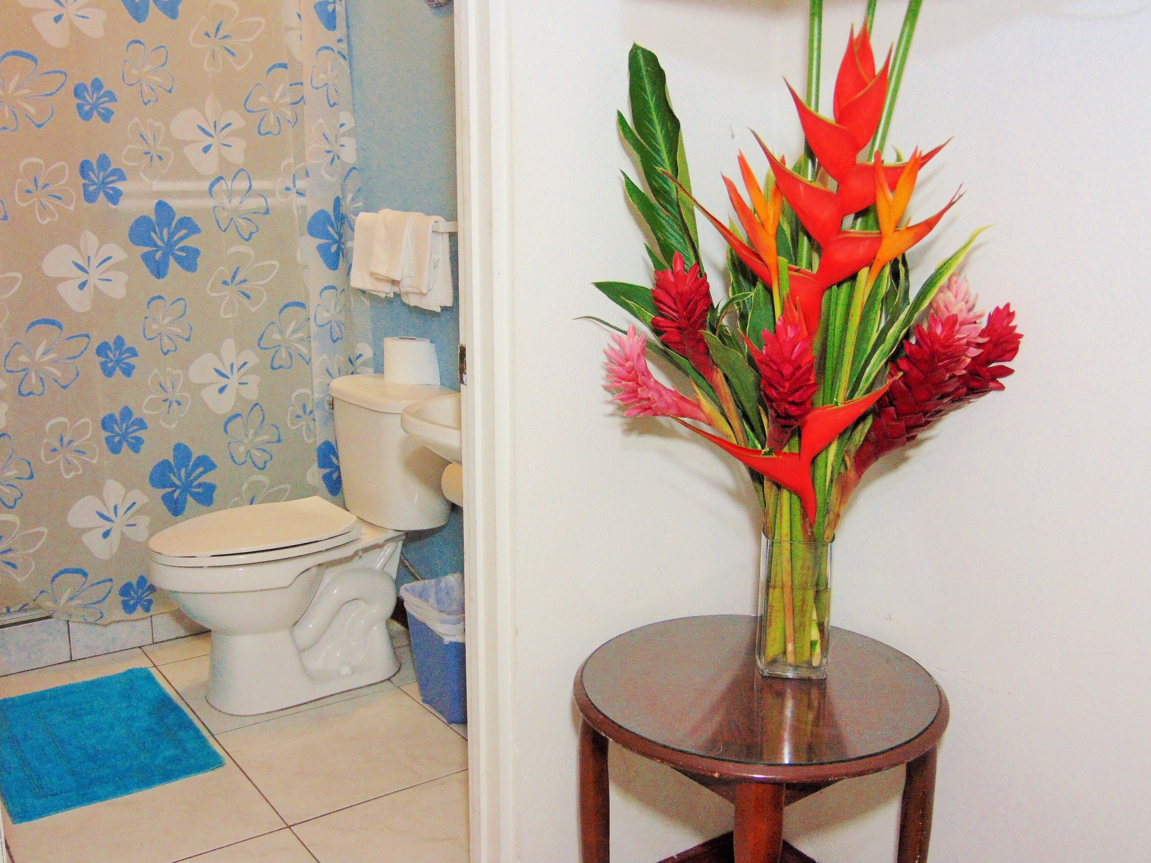 A bathroom with a toilet, a blue mat, and a vase with red flowers on a small table.