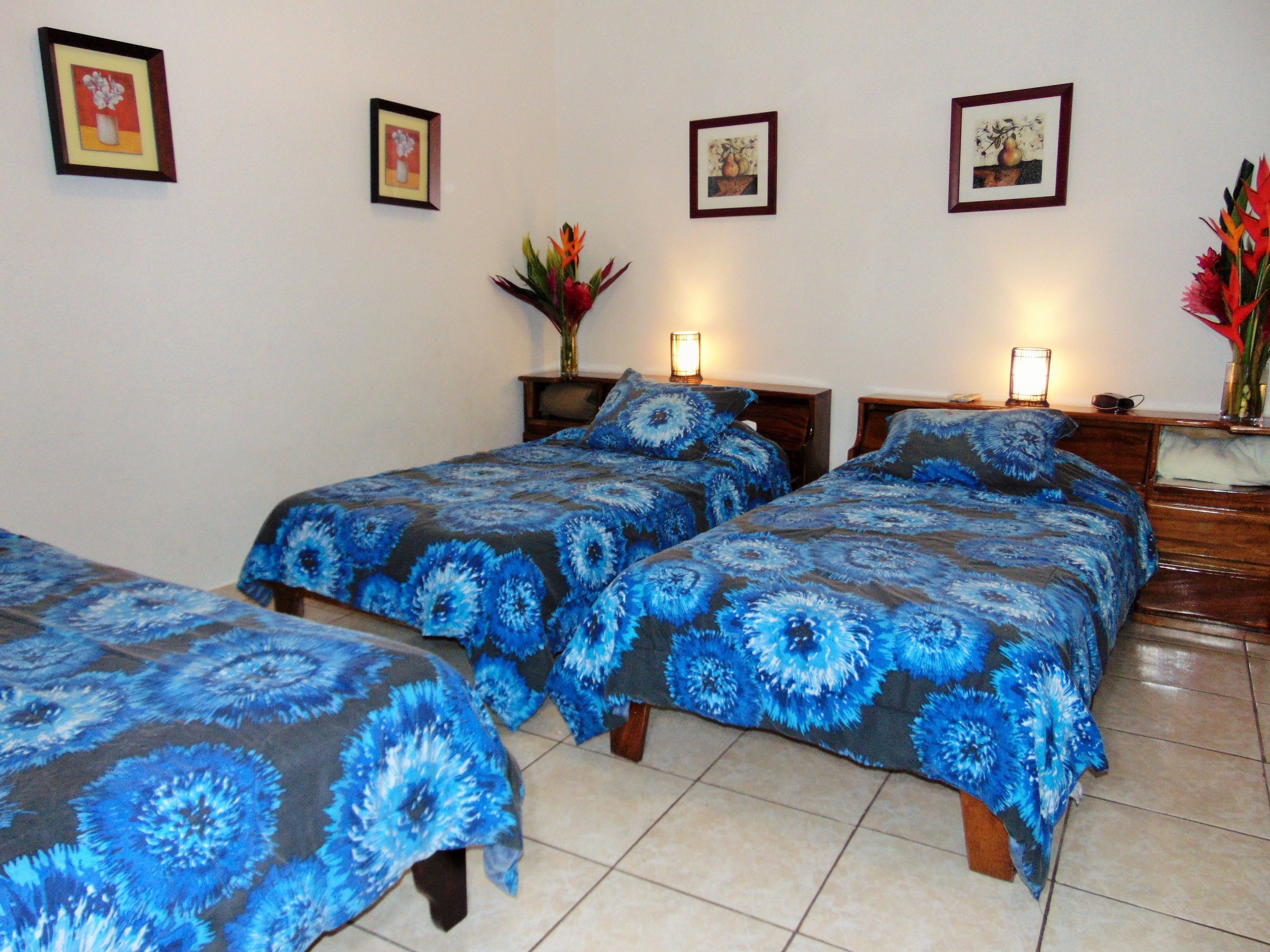 A hotel room with two single beds, a nightstand, a bedside table, a vase with flowers, and framed artwork on the wall.