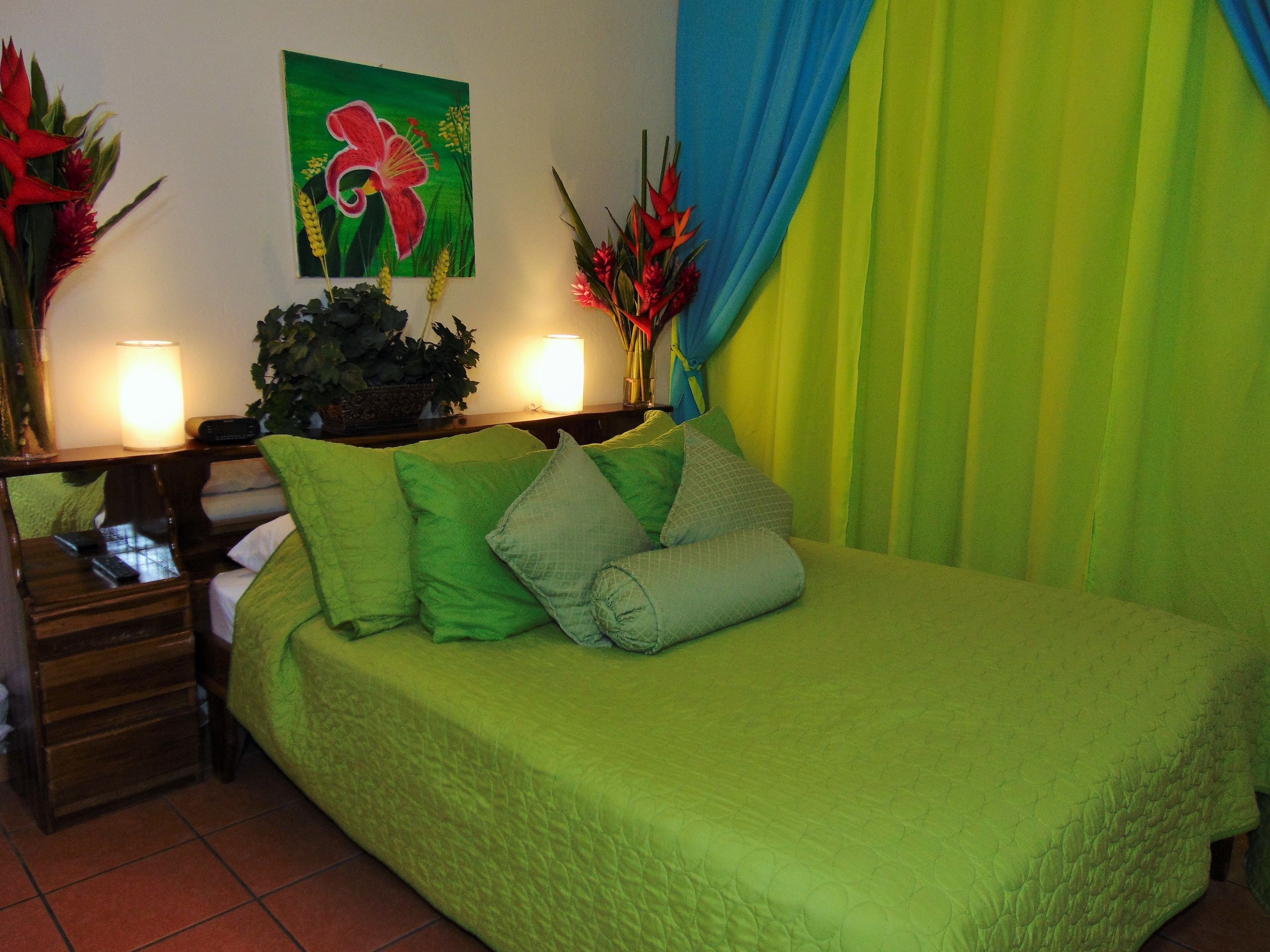 A room with a green bed, two bedside tables with lamps, a painting, and a curtain.