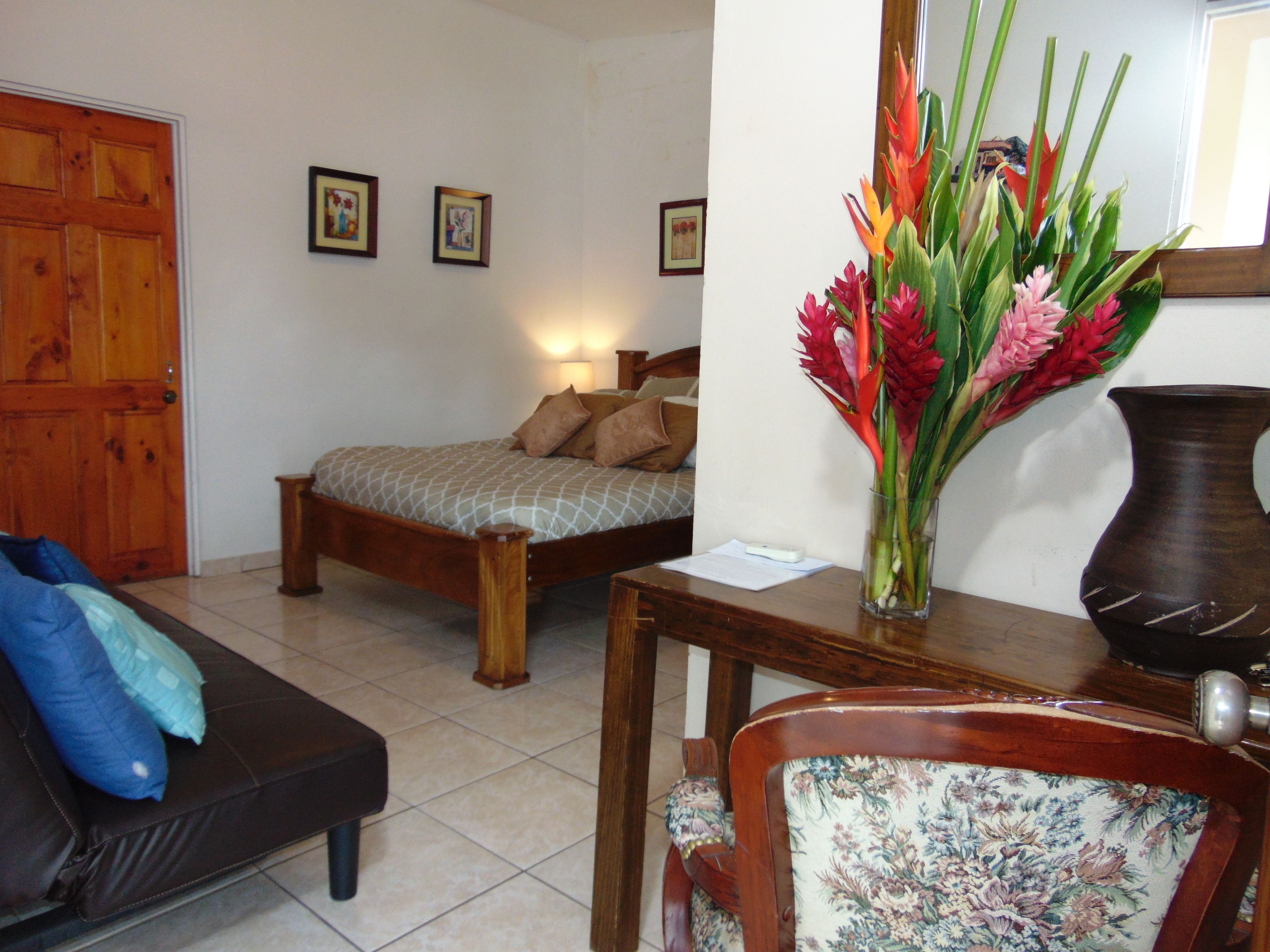 A bedroom with a bed, a wooden door, a bedside table with a vase of flowers, a chair with floral upholstery, and a framed picture on the wall.
