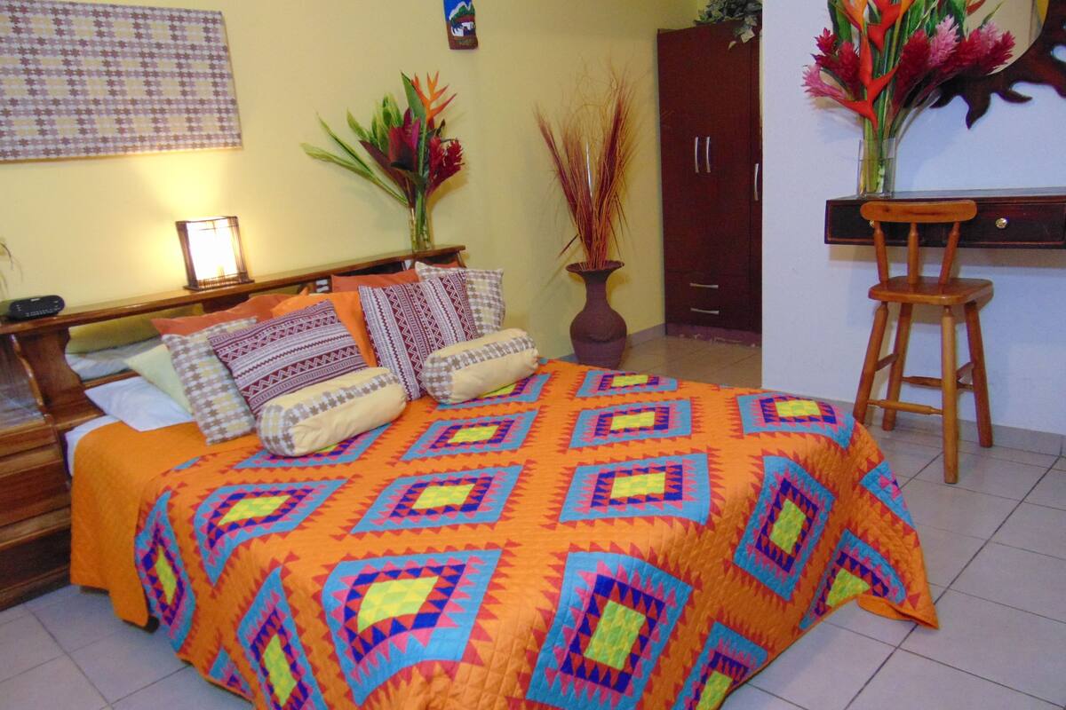 A bed with a colorful quilt, a wooden nightstand, a bedside table with a lamp, a wooden wardrobe, and a chair.