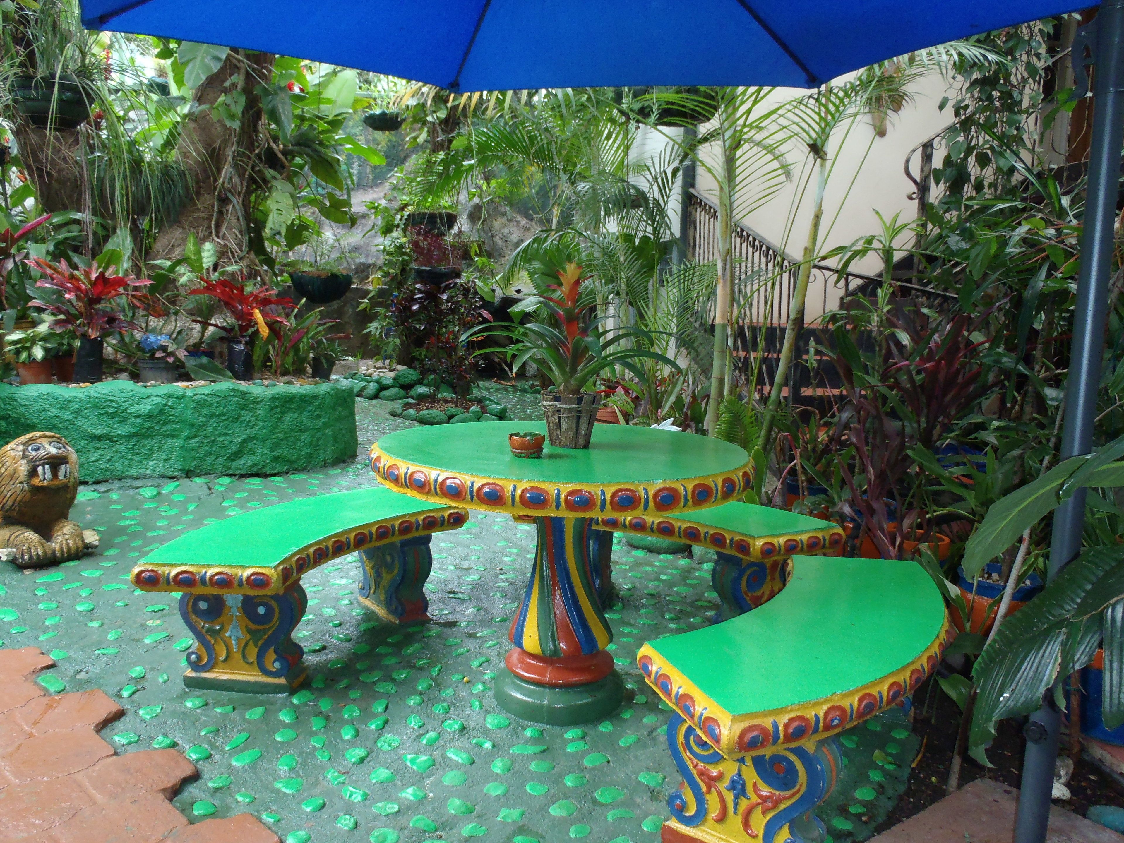Un espacio al aire libre con ambiente tropical, con una mesa redonda y bancos, rodeado de exuberante vegetación y plantas en macetas.