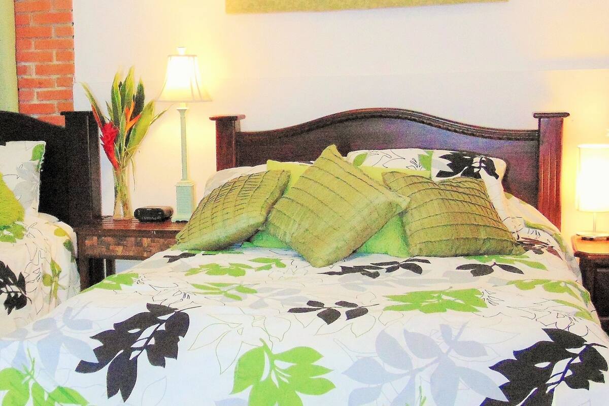 A hotel room with two beds, each with floral bedding and multiple pillows. There are bedside tables with lamps and decorative vases.