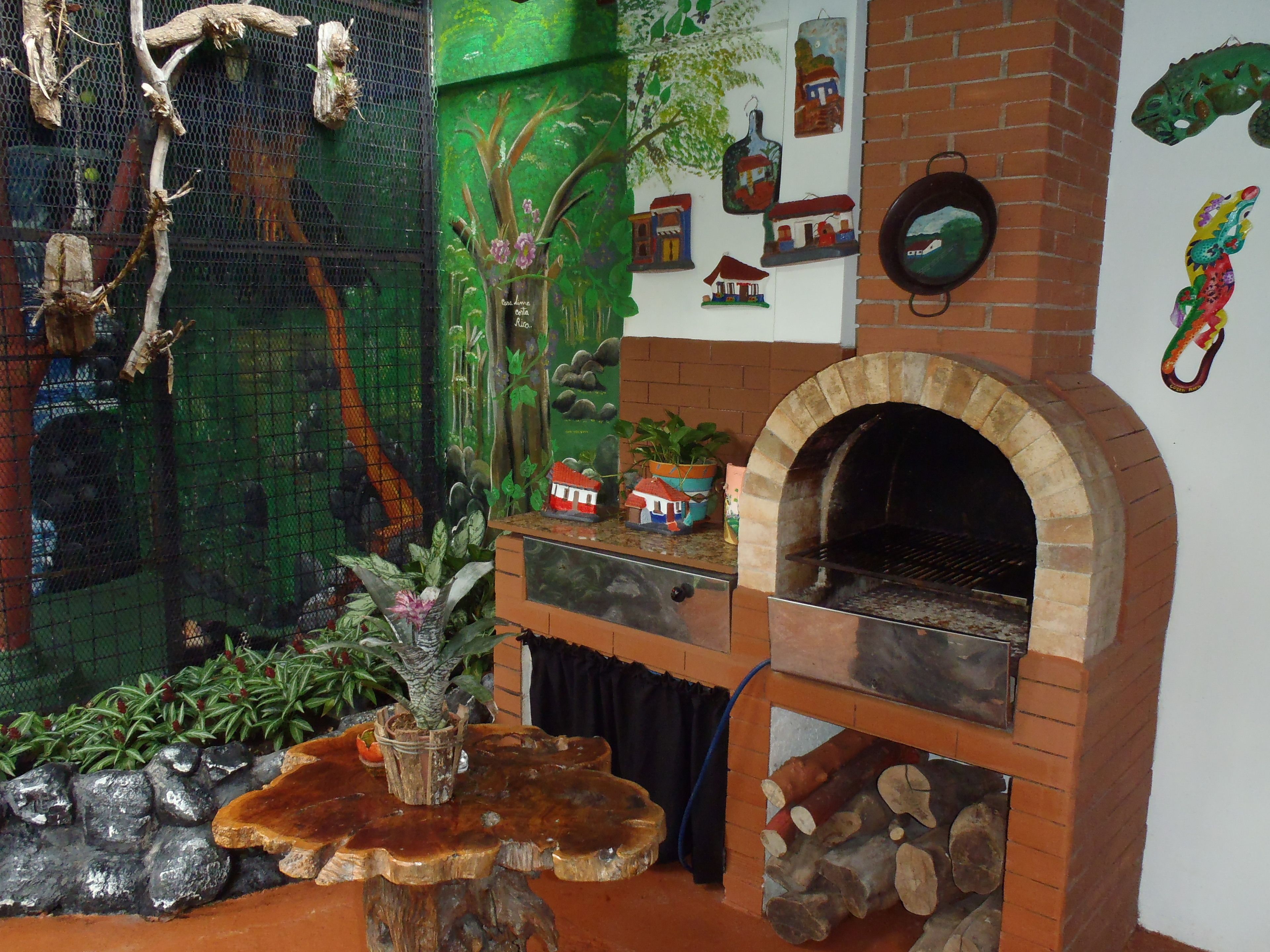 Un interior rústico con chimenea de ladrillo, una mesa de madera y diversos objetos decorativos, incluyendo una jaula de pájaros y adornos en la pared.