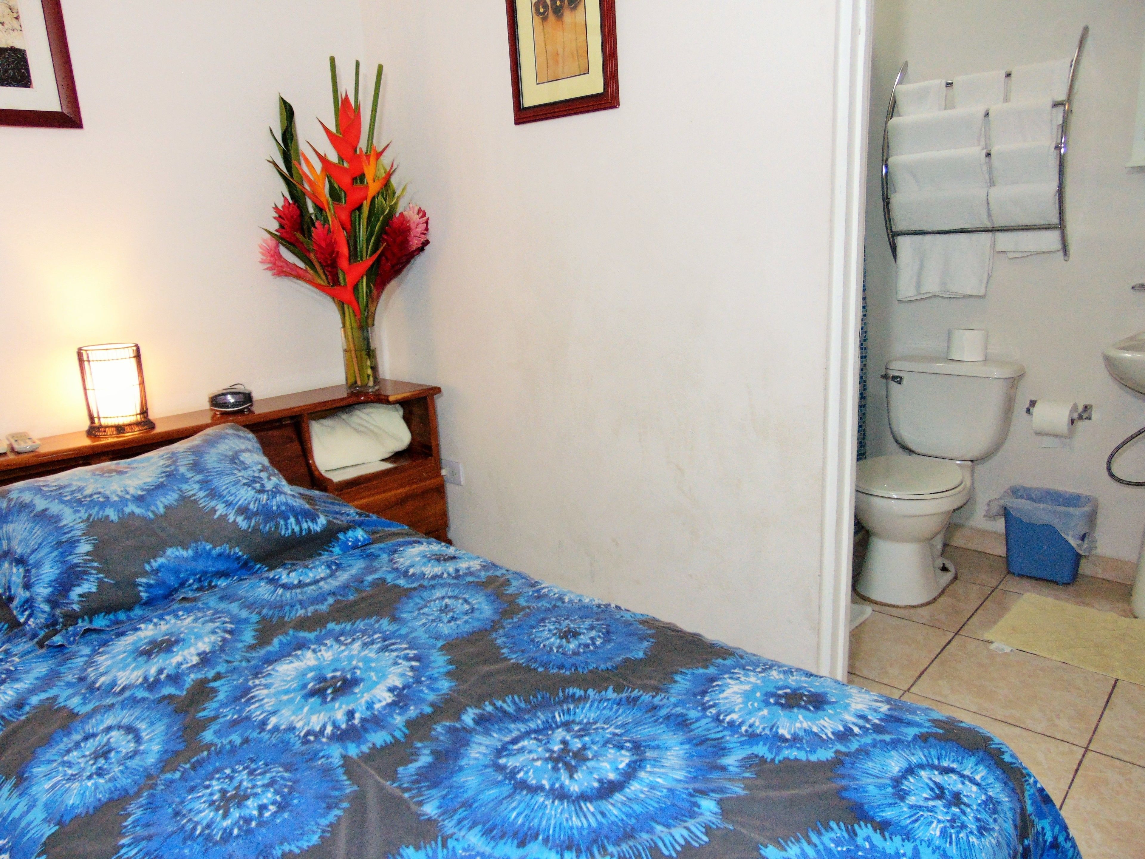 A bedroom with a bed, a nightstand, a lamp, a vase with flowers, and a bathroom with a toilet and sink.