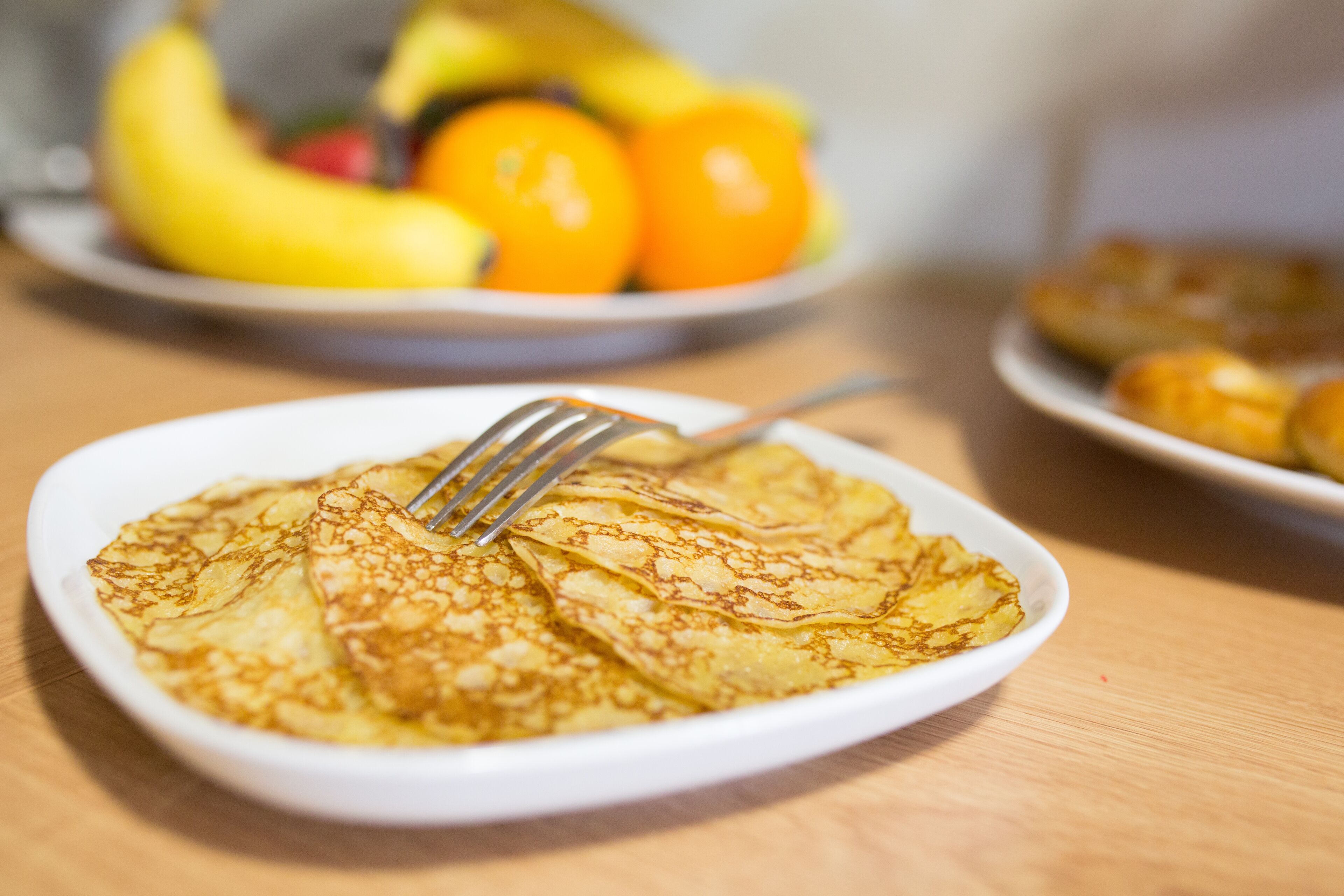 A plate of crepes with a fork on it, a bowl of fruit in the background, and another plate with pastries.