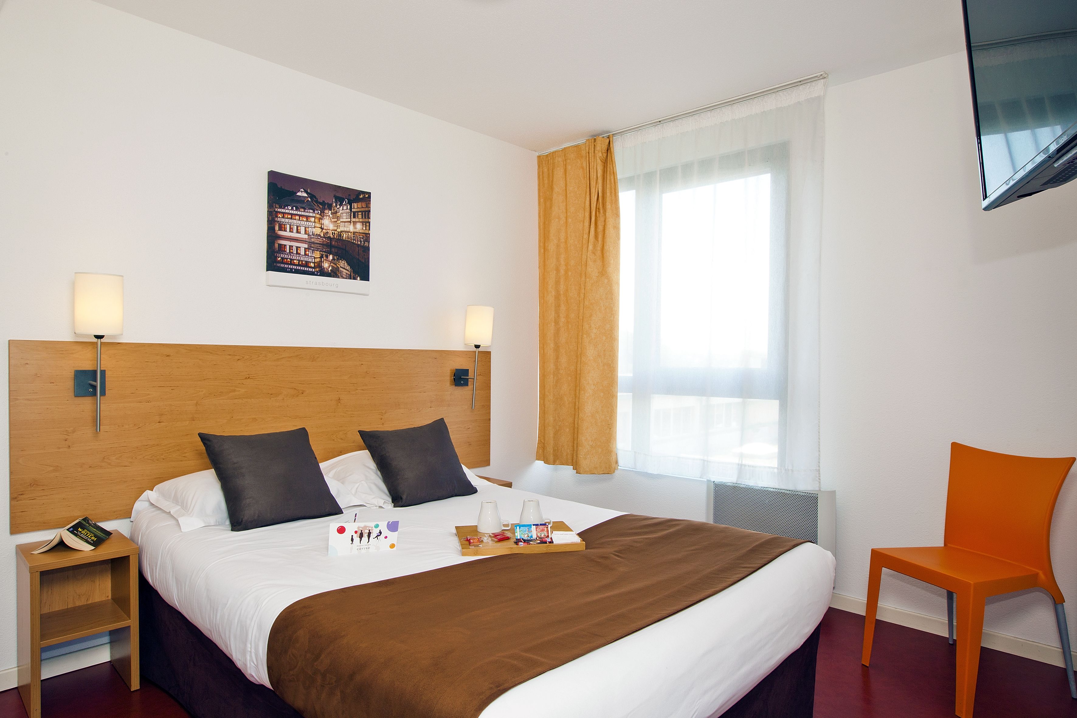 A hotel room with a large bed, a wooden headboard, two bedside tables, a nightstand, a window with curtains, and a television mounted on the wall.