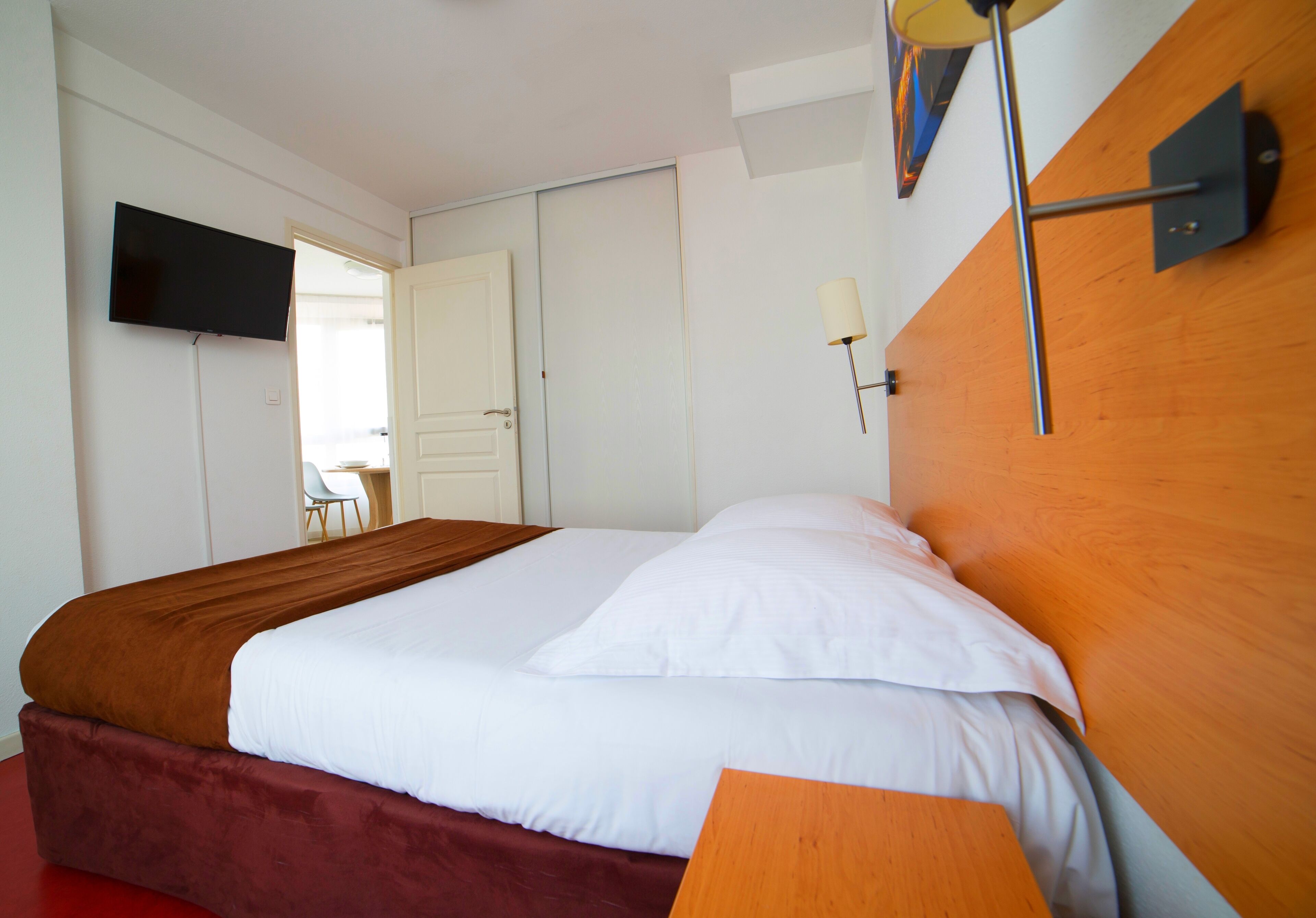 Habitación de hotel con cama, televisión montada en la pared, lámpara de noche y puerta que da a otra habitación.