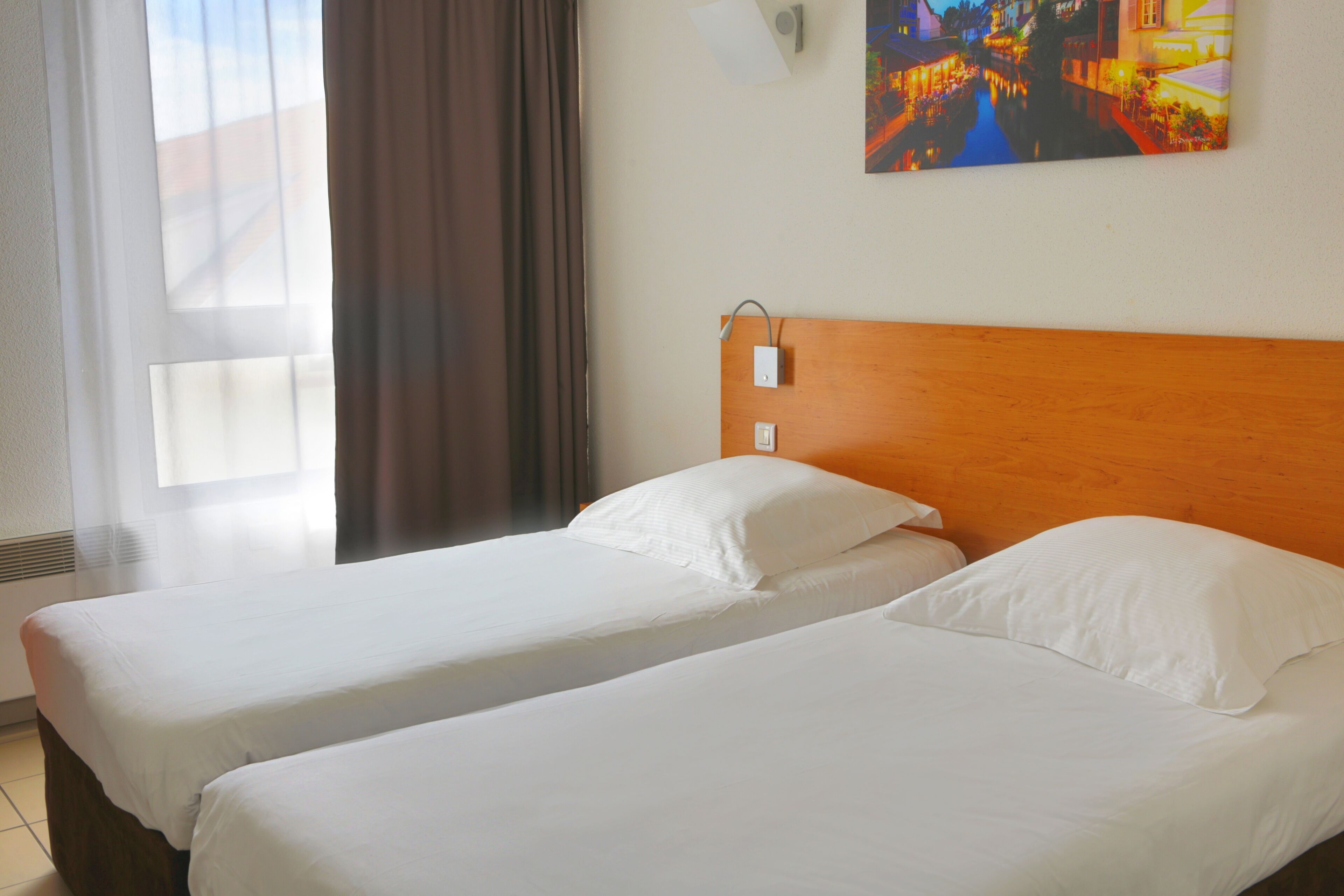 A hotel room with two single beds, a wooden headboard, a window with curtains, and a painting on the wall.