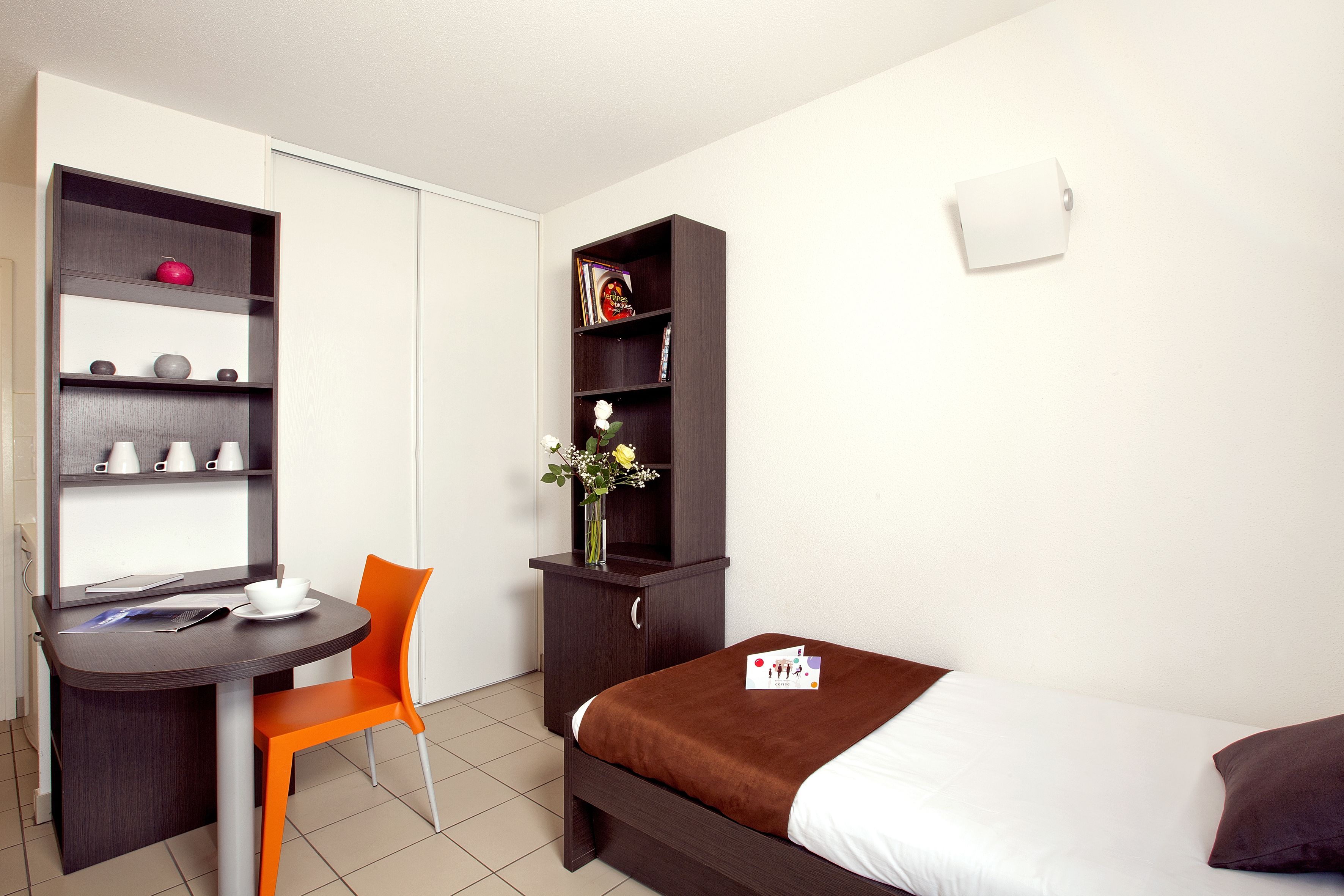 A compact hotel room with a bed, a desk with a chair, a bookshelf, and a small cabinet.