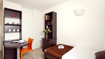 A compact hotel room with a bed, a desk with a chair, a bookshelf, and a small cabinet.