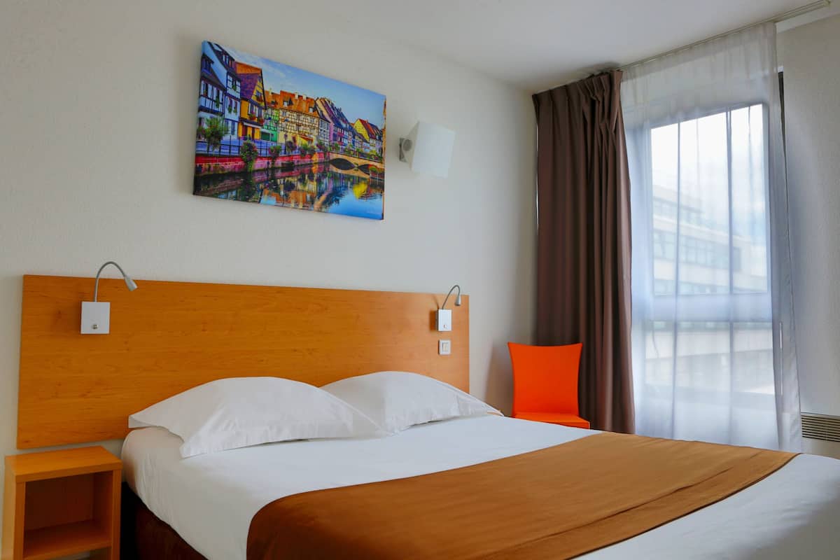 Une chambre d’hôtel avec une tête de lit en bois, un grand lit avec une literie blanche et marron, un tableau coloré accroché au mur, une table de chevet, une chaise orange et une fenêtre avec des rideaux.