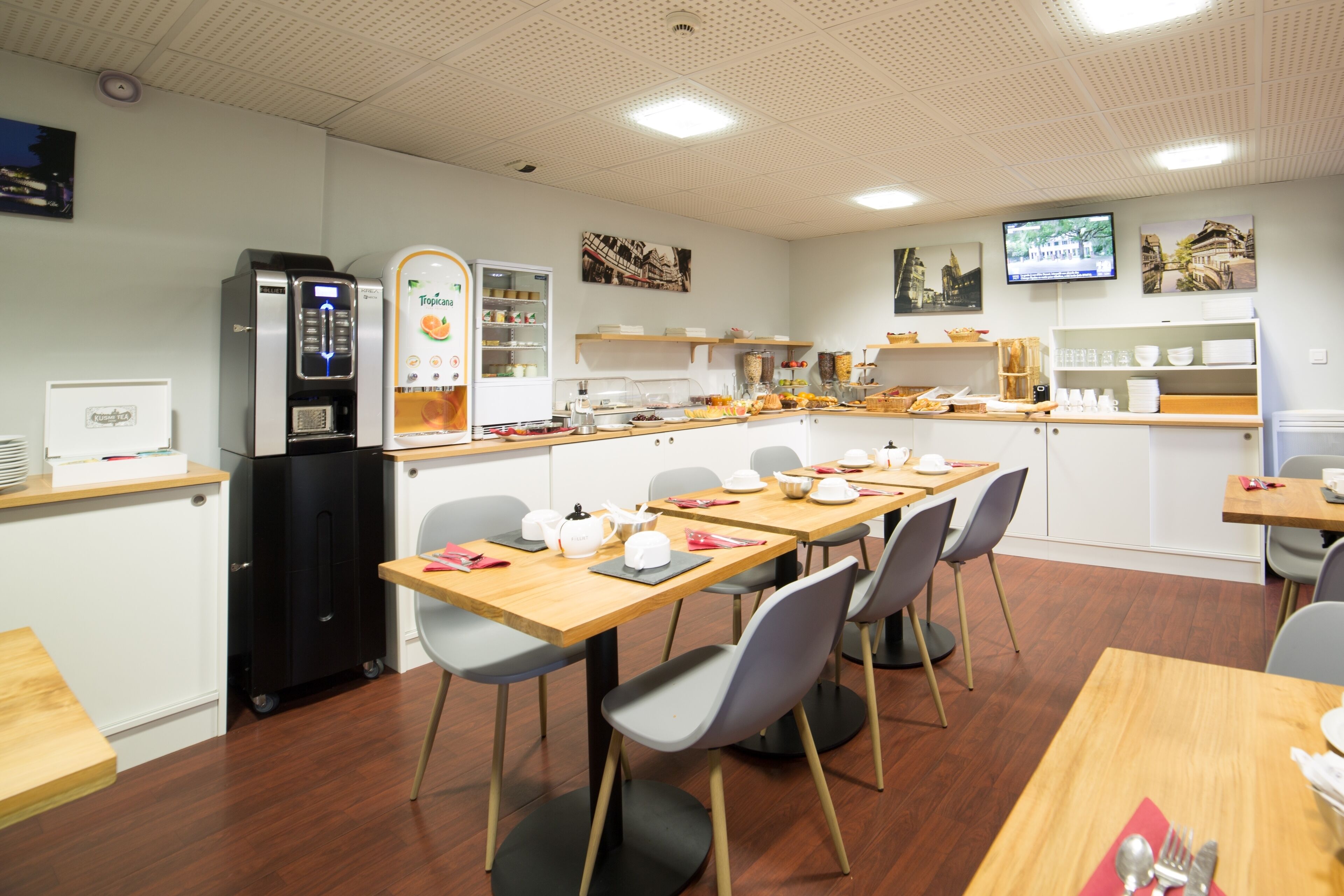 A modern dining area with tables and chairs, a refrigerator, a coffee machine, and a television mounted on the wall.