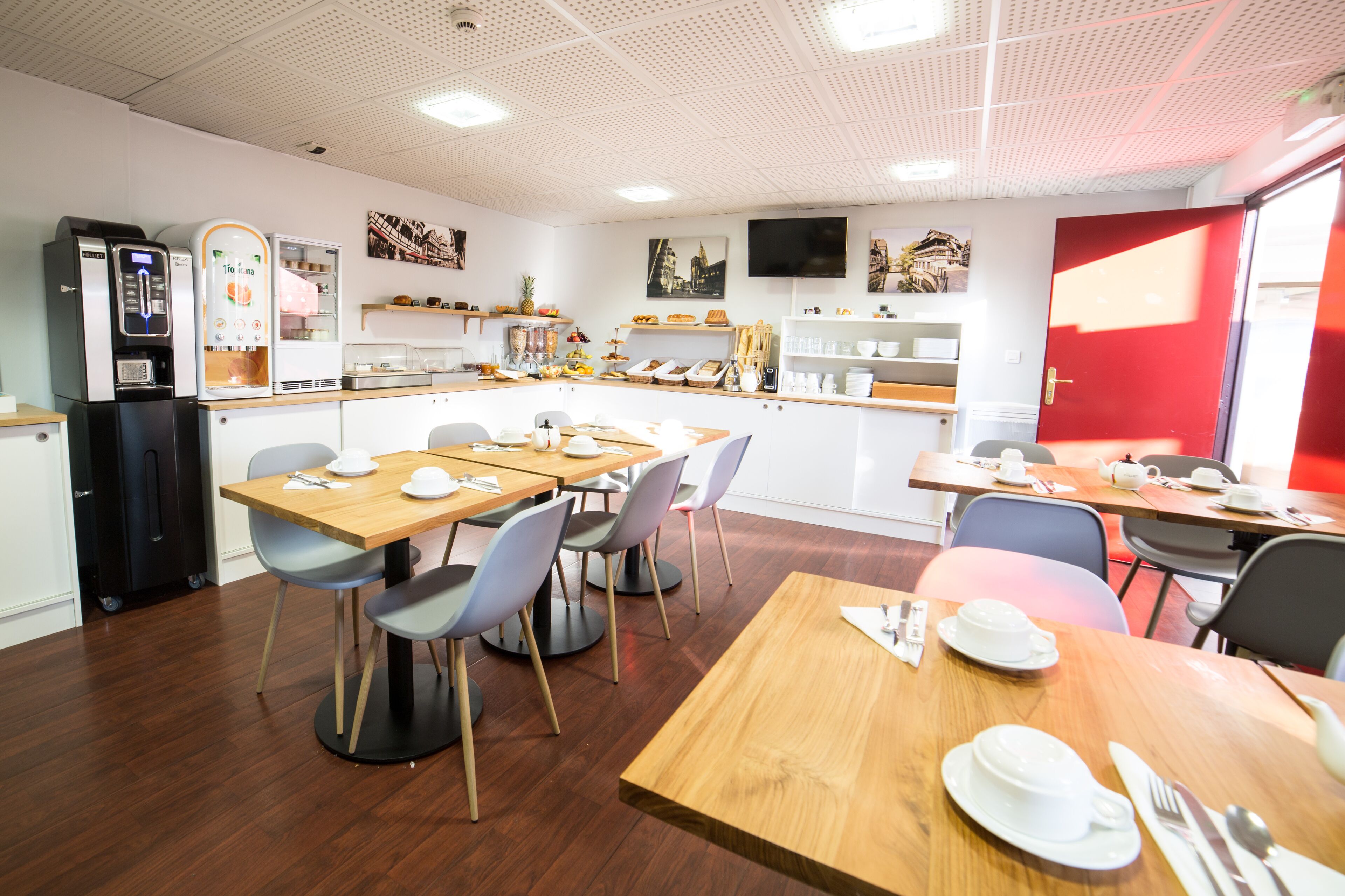 A modern dining area with wooden tables and chairs, a refrigerator, a coffee machine, and various food items on the counter.