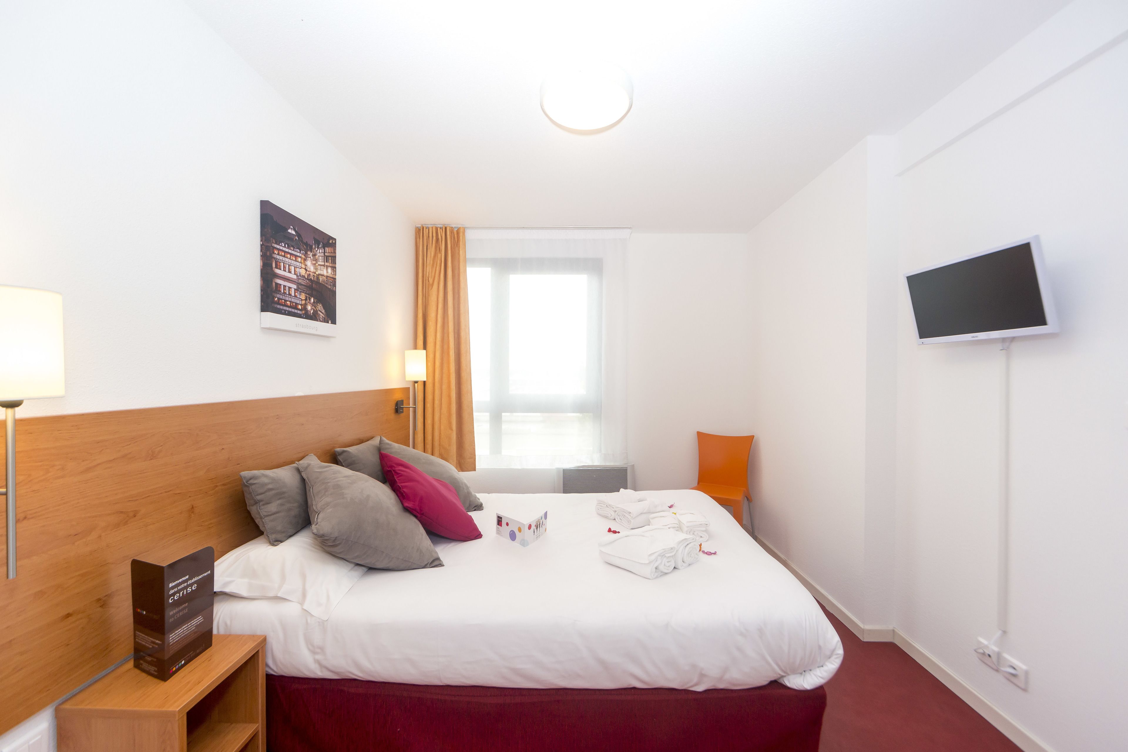 A hotel room with a neatly made bed, a flat-screen TV mounted on the wall, and a window with curtains.