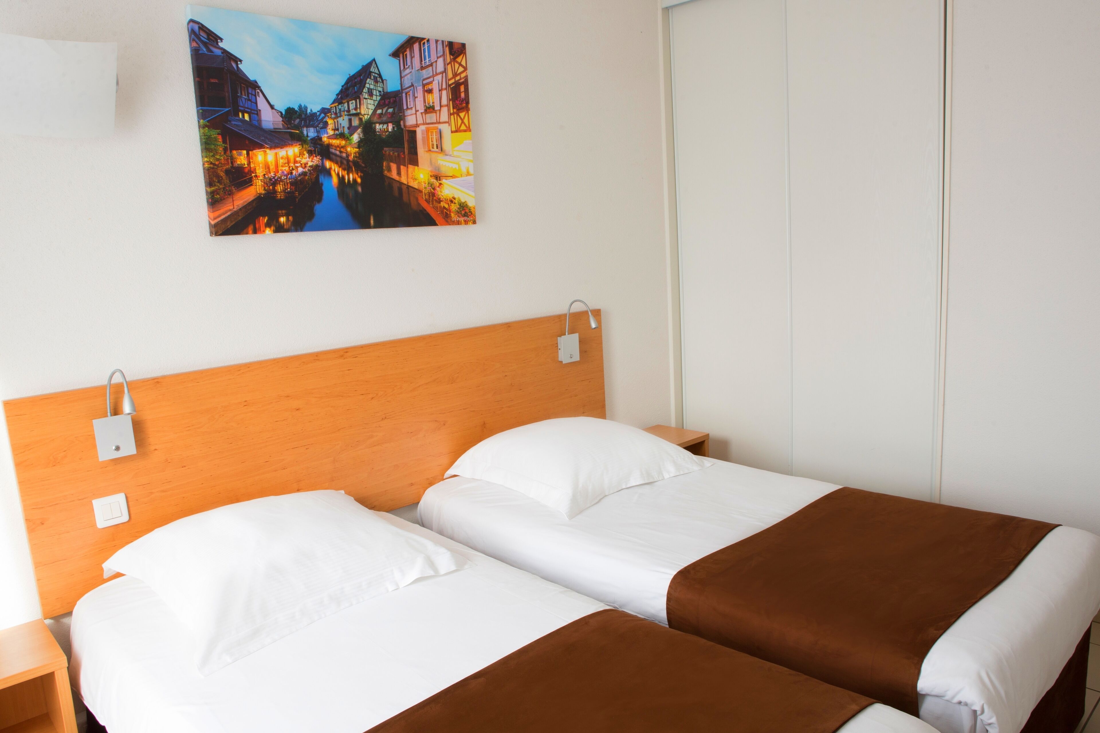 Habitación de hotel con cabecera de madera, dos camas con ropa de cama blanca y marrón, una mesita de noche, un enchufe en la pared y una imagen de una escena de canal.
